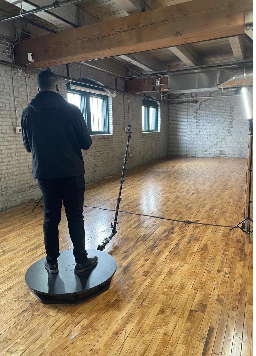 A man stands on a rotating platform in a studio with wooden floors, metal pipes, brick walls, and windows, as he undergoes a balance test or assessment with professional lighting and equipment.