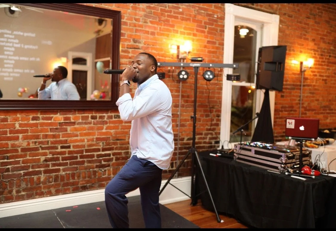 A man singing into a microphone at an indoor party or event with DJ equipment and a large mirror on a brick wall behind him.
