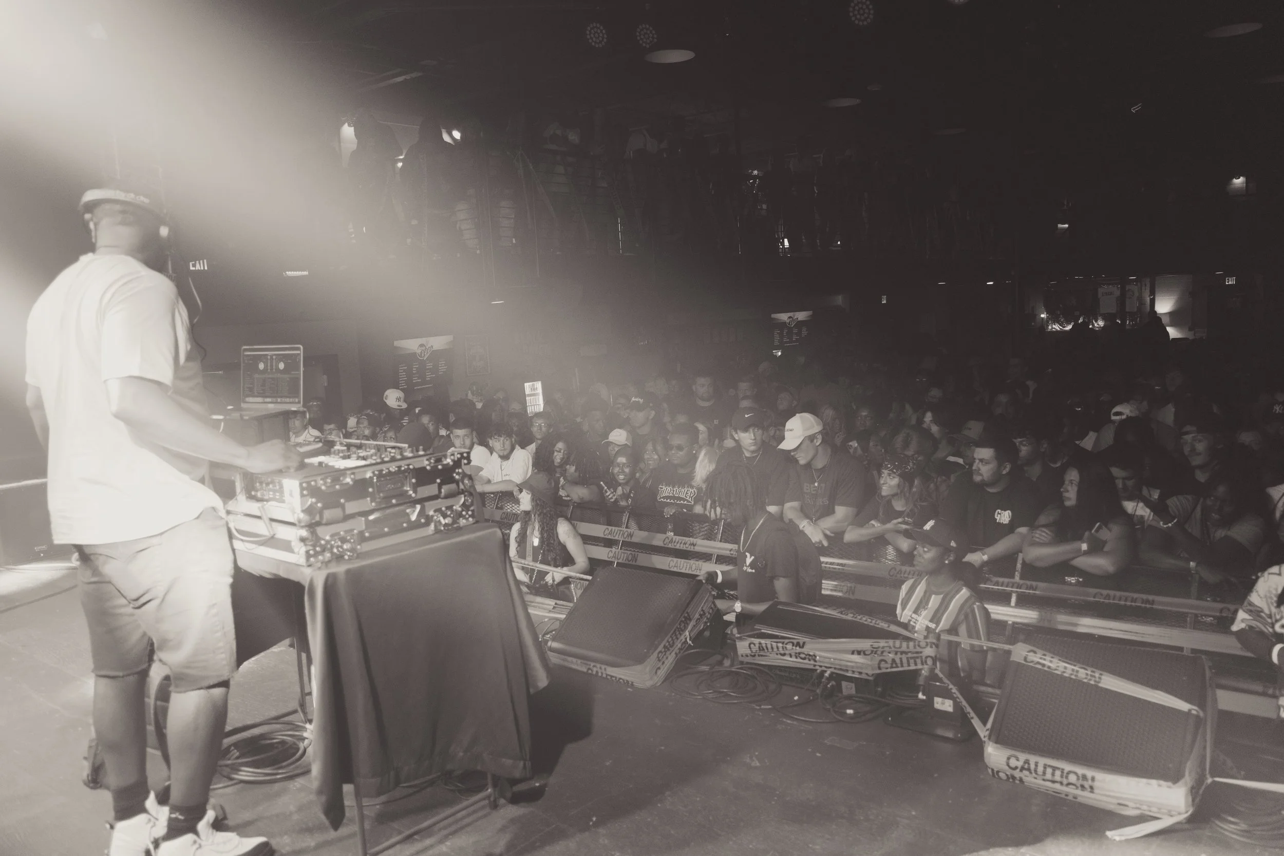 A DJ in a white shirt and shorts operating turntables and audio equipment in front of a crowded dance floor at a nightclub or concert venue. The crowd is facing the DJ and enjoying the performance. Concert DJ