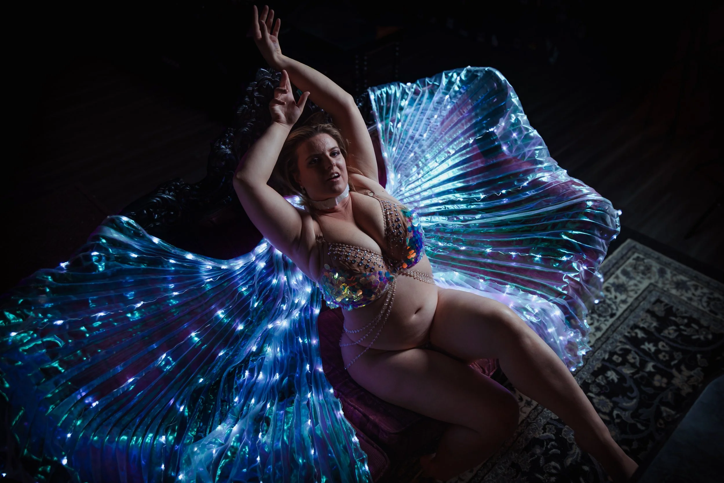 A woman lying on a dark floor with her arms raised, wearing an iridescent, illuminated costume with lights radiating outwards.