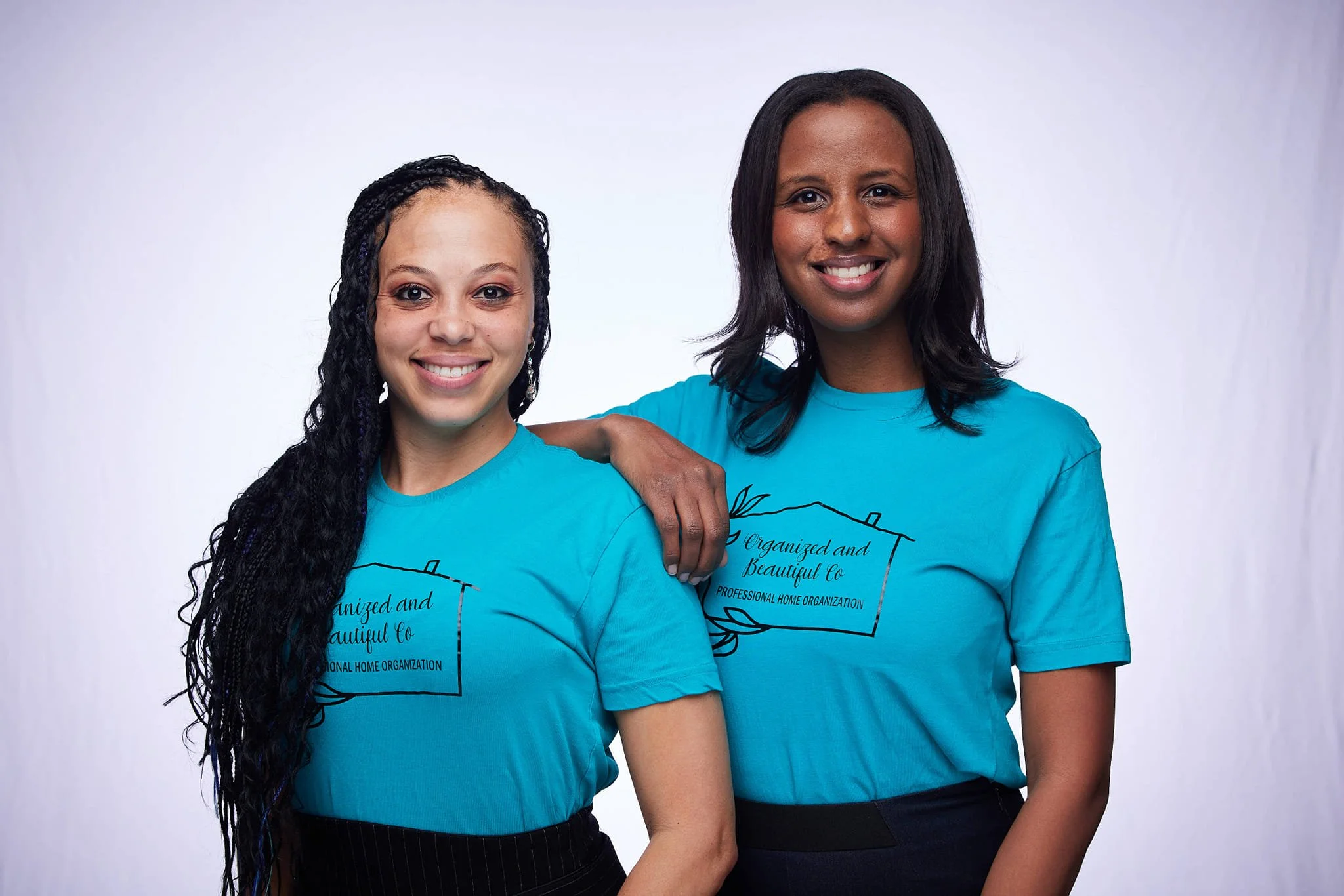 Two women wearing matching blue T-shirts with a logo and text that includes 'Organized and Beautiful Co' and 'Professional Home Organization,' standing close together and smiling in front of a plain white background.