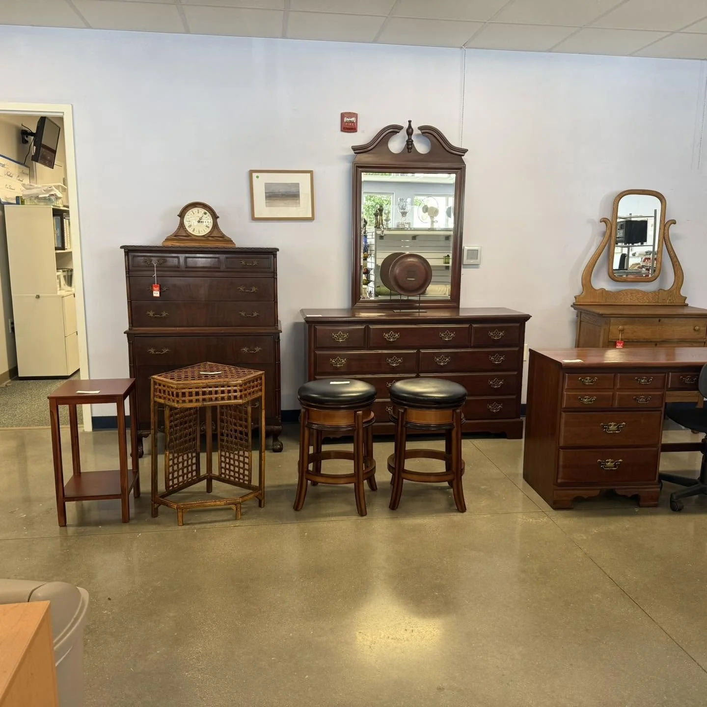 A furniture display with five wooden furniture pieces including a tall dresser, a dresser with a mirror, a small side table, and two stools, arranged against a white wall in a store or showroom.