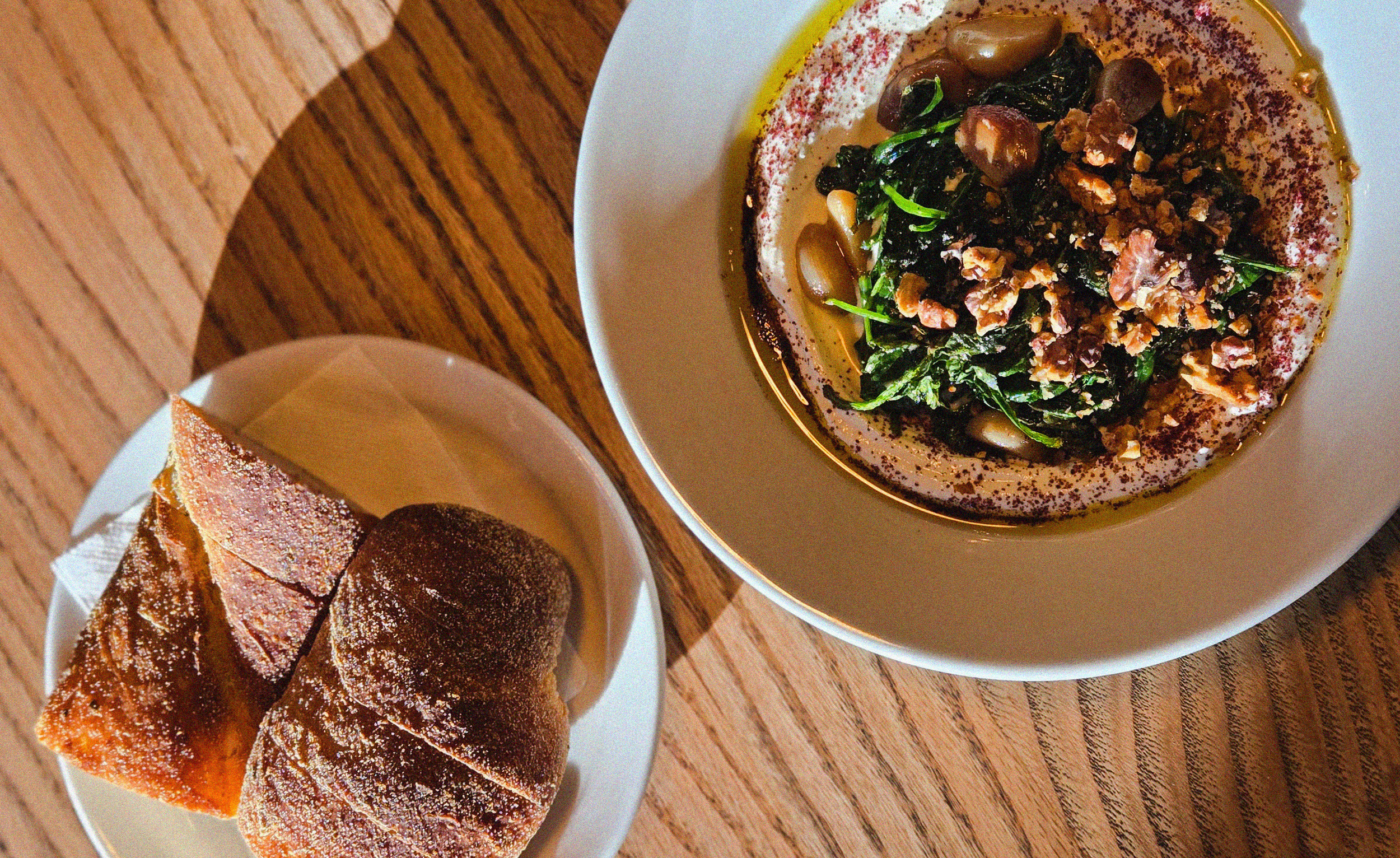 A plate of hummus topped with herbs, sumac, chopped walnuts, and olive oil, served on a white plate. Next to it, a small white bowl contains three pieces of baklava, a layered pastry with a sugar glaze, on a wooden table.