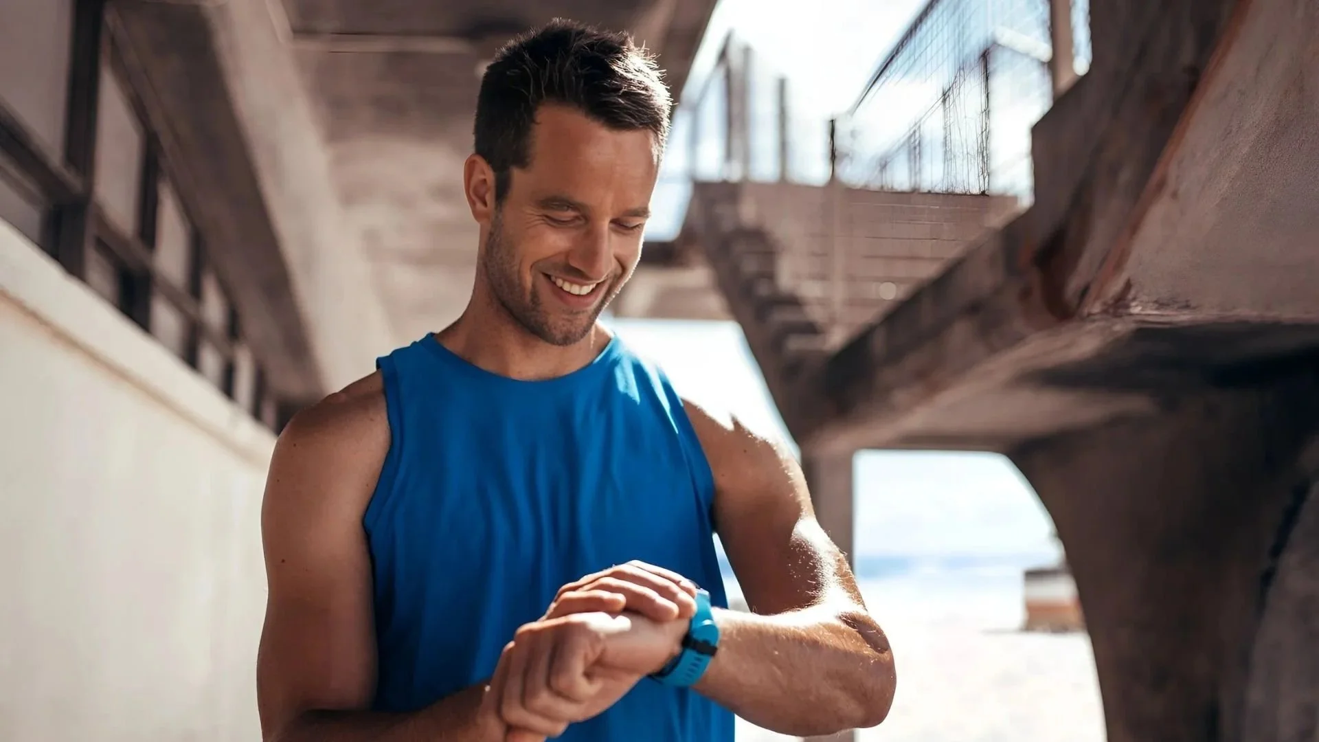 A man in a blue athletic tank top smiling and looking at his smartwatch while standing outdoors under a wooden structure near a beach.