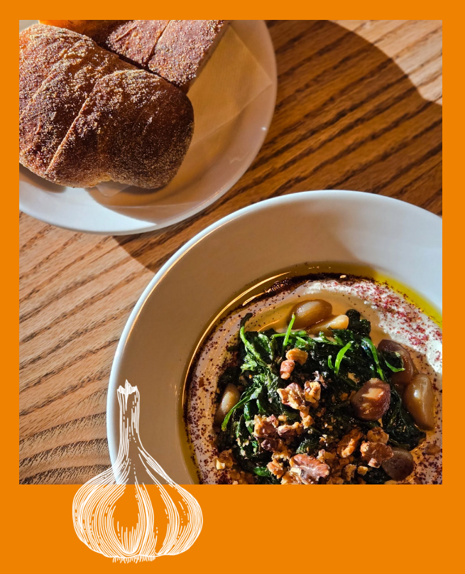 A bowl of creamy hummus topped with greens, pine nuts, and walnuts, next to a plate of toasted bread on a wooden table.