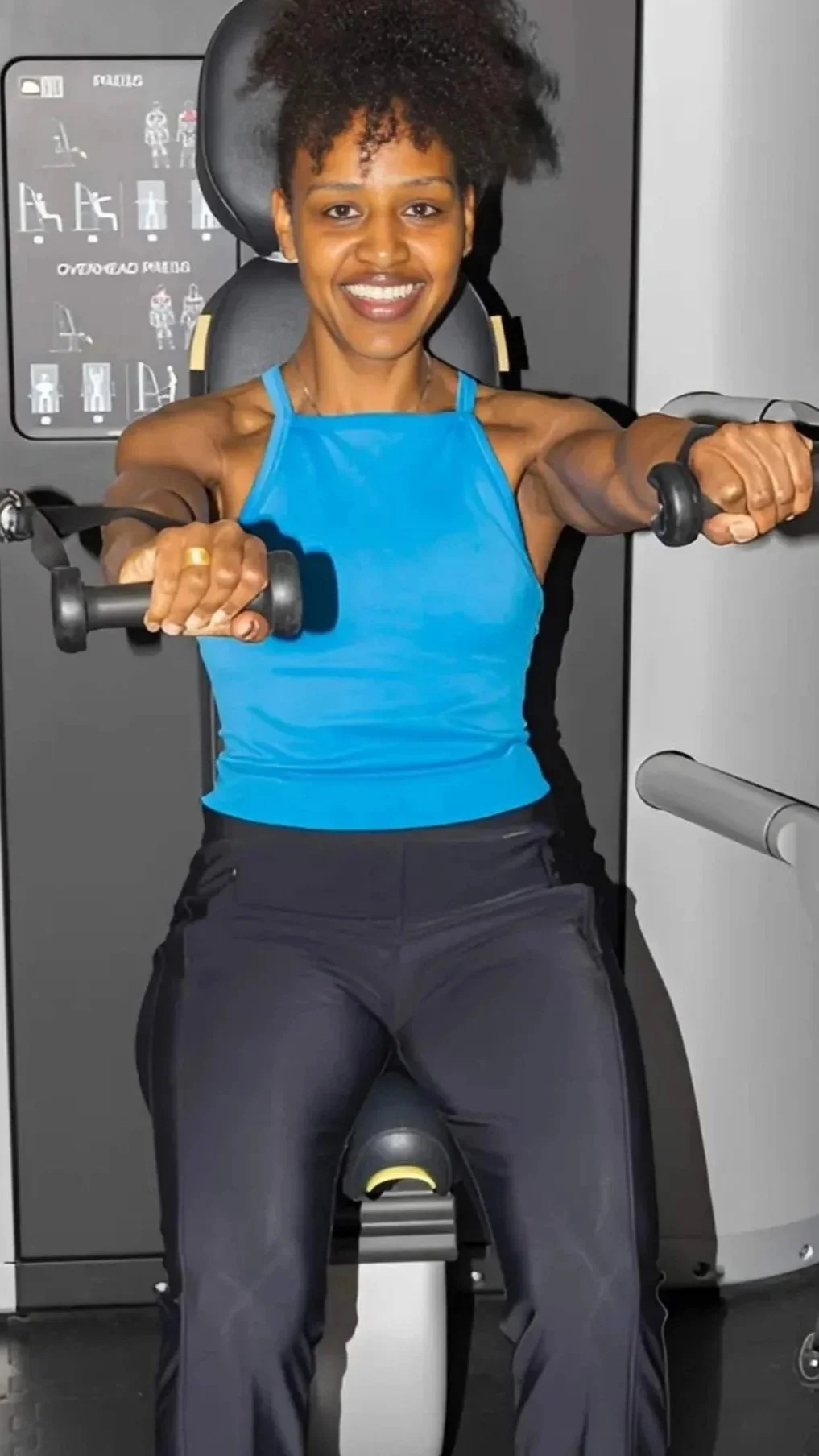 Menopause Coach, Aman on a fitness machine, smiling, wearing a blue tank top and black athletic pants, exercising with hand weights.