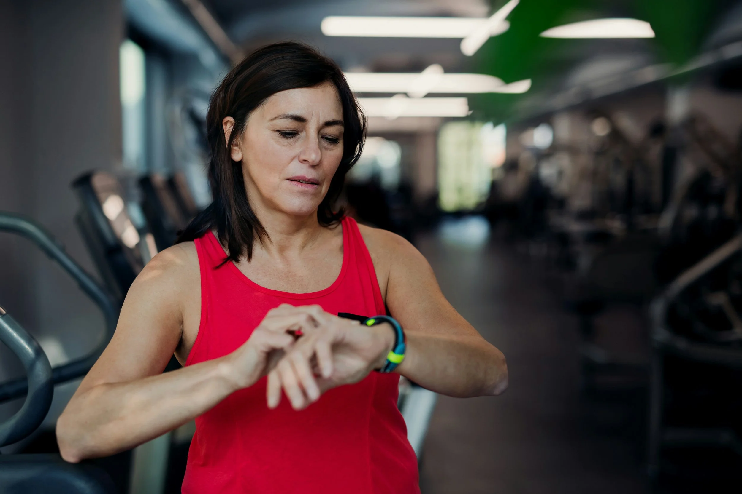 Woman in a red tank top checking her smartwatch in a gym.