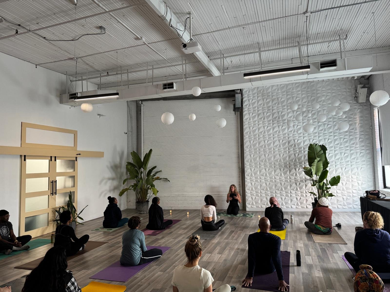 A group of people participating in a yoga class in a spacious, modern studio with white walls, wooden floors, large green potted plants, and decorative white paper lanterns hanging from the ceiling. The instructor is sitting at the front, speaking to the class.