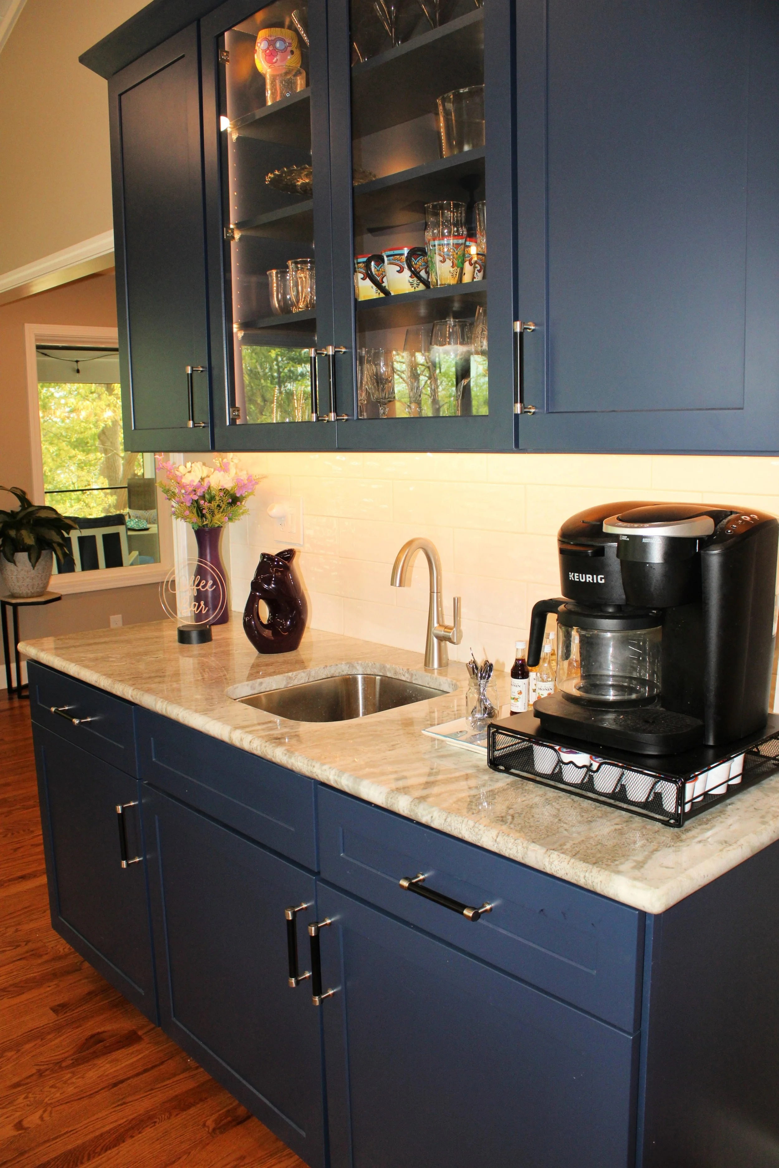 Kitchen with navy blue cabinets, a marble countertop, a sink, and a Keurig coffee maker. Open glass cabinet with decorated glasses and mugs. Decor items including a purple vase with flowers and a dark ceramic bird figure.