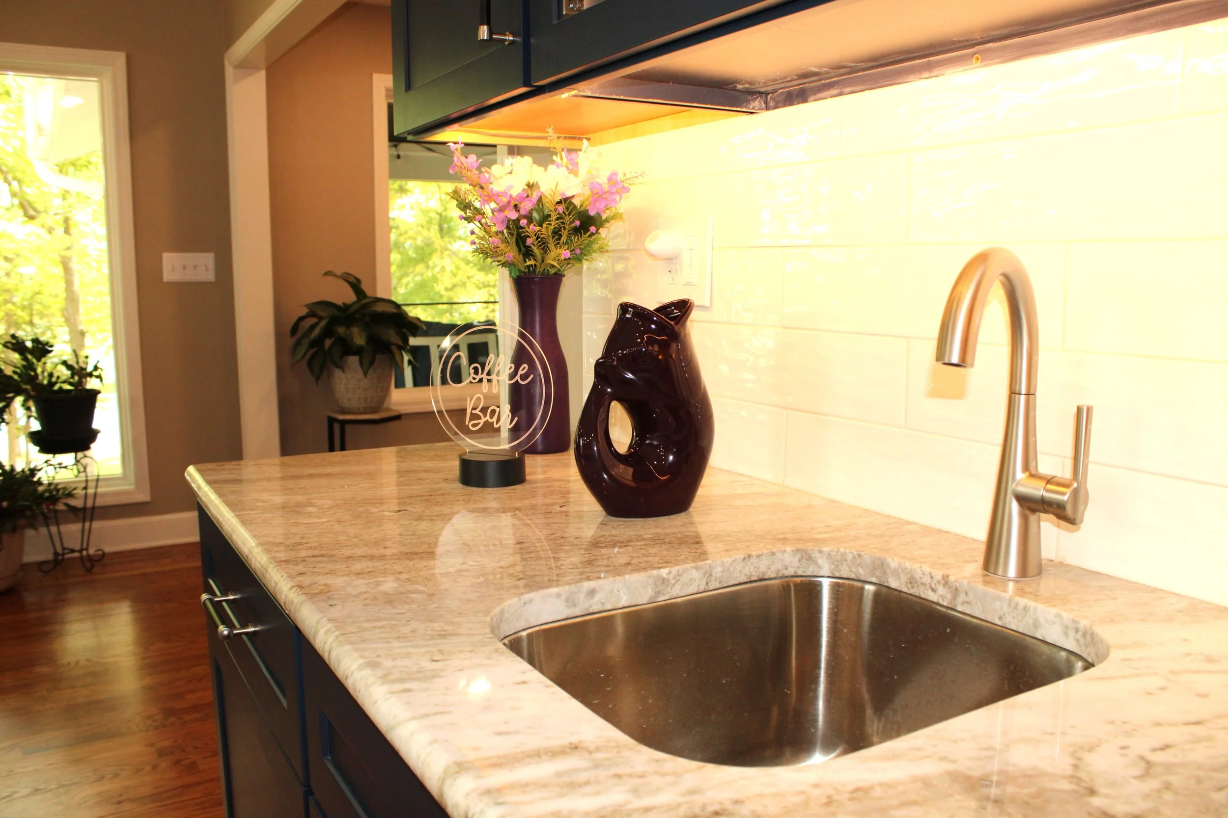 Close-up of a kitchen countertop with a sink, a dark ceramic vase with flowers, a black decorative sculpture, and a lighted sign that reads 'Coffee Bar'.