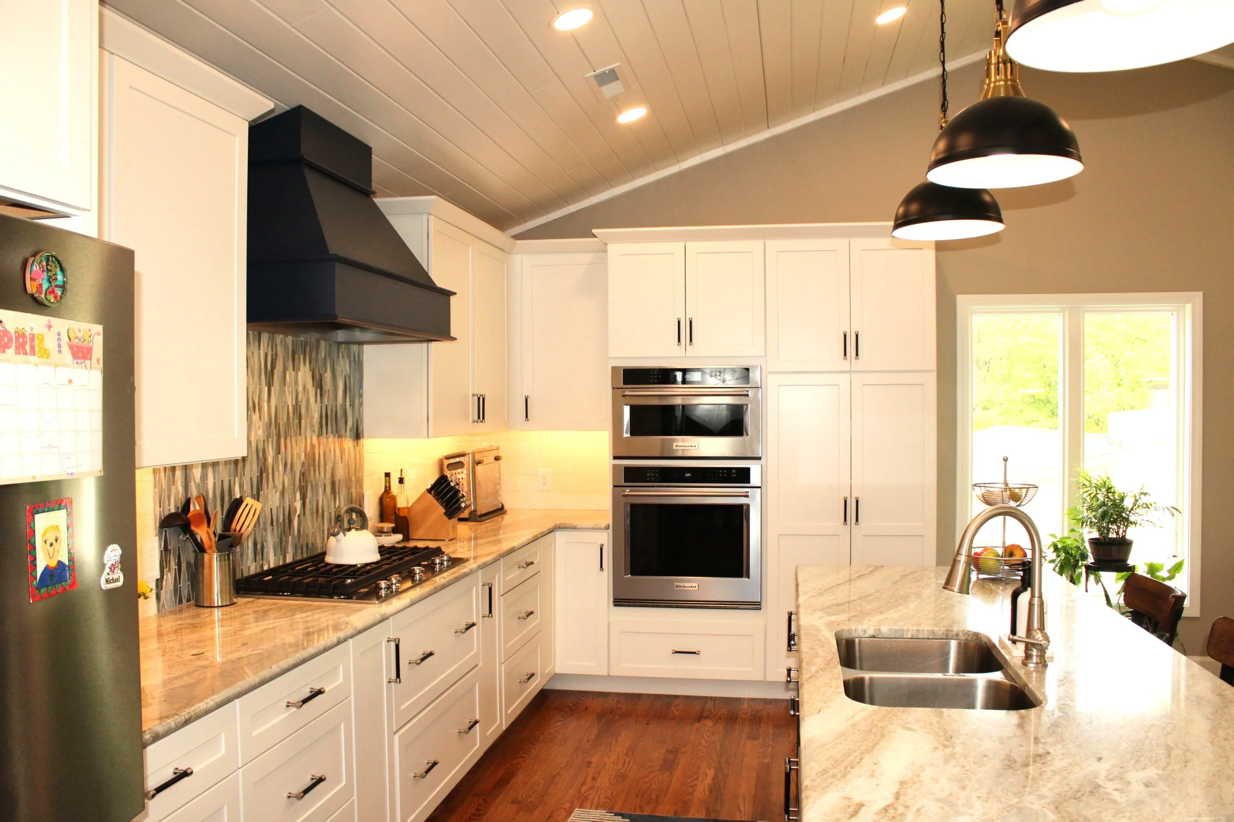 Modern kitchen with white cabinets, beige marble countertops, stainless steel appliances including a double oven, a black range hood, and a mosaic tile backsplash. Large window provides natural light, and black pendant lights hang from the ceiling.