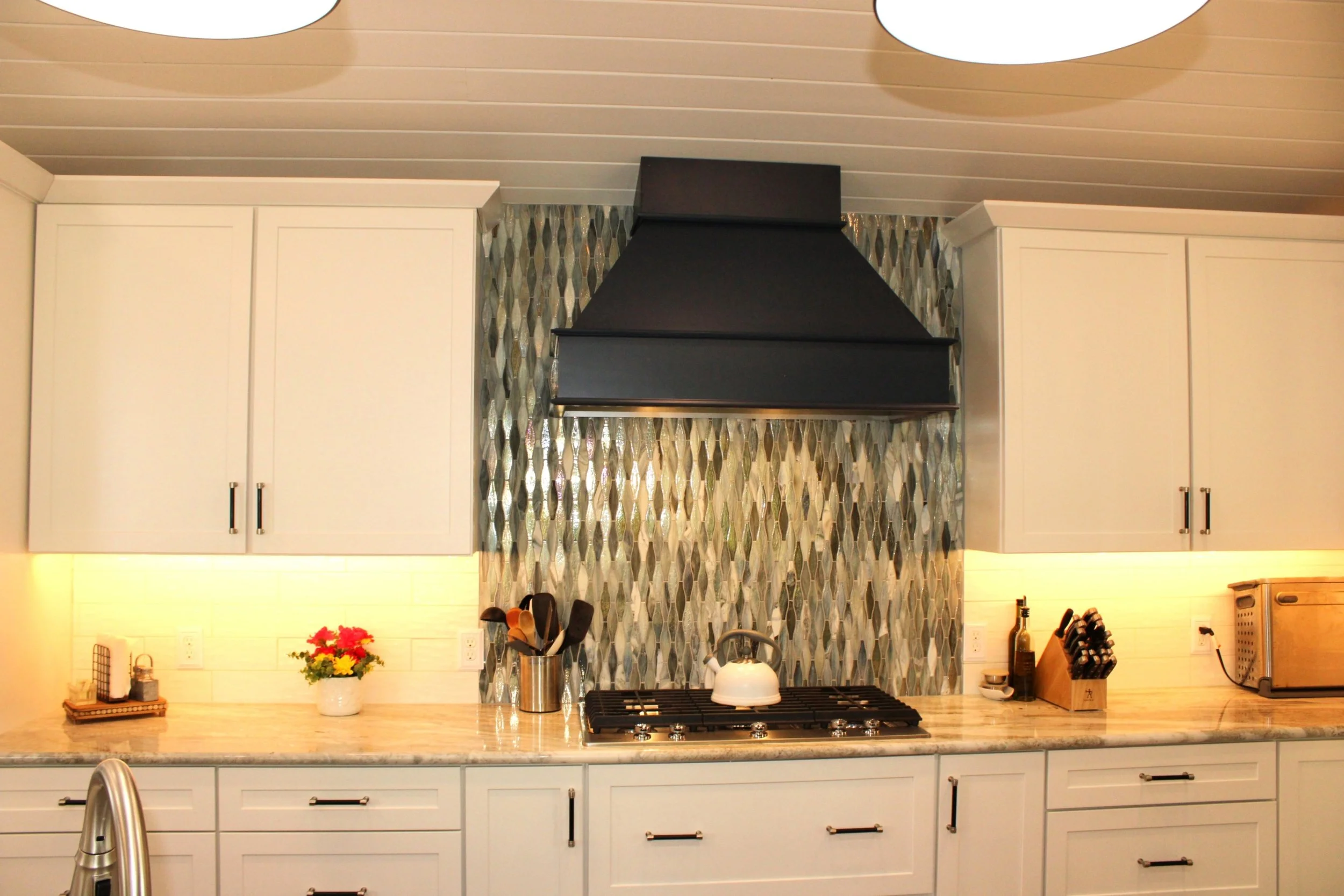 A modern kitchen with white cabinets, a black range hood, patterned tile backsplash, and a marble countertop with kitchen utensils, a potted plant with flowers, and various small appliances and containers.