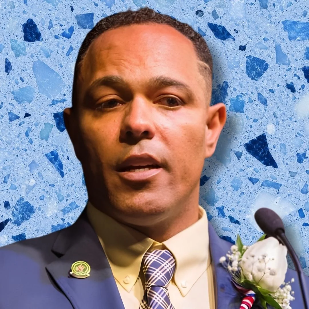 A man in a suit speaking at a podium with a microphone, wearing a flower boutonniere, against a blue terrazzo background.