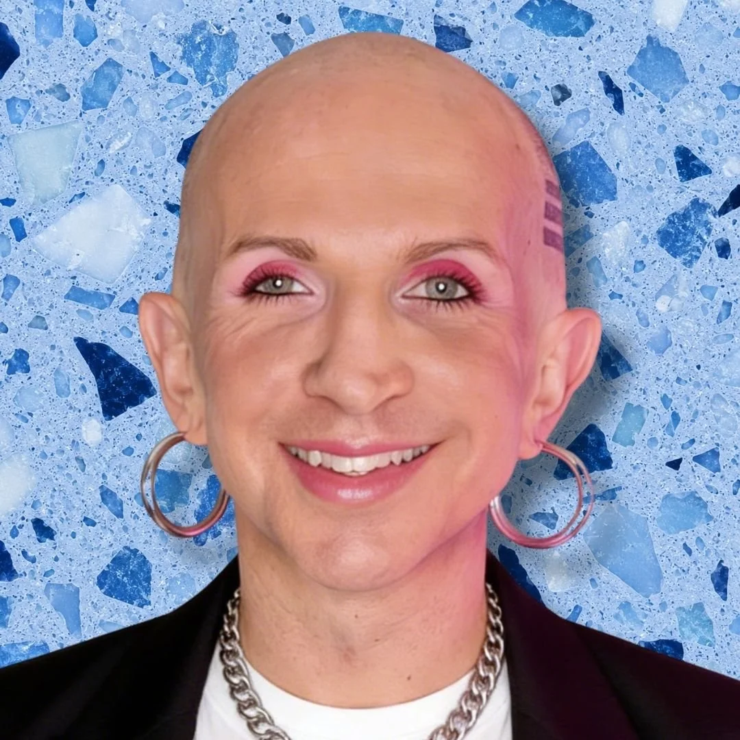 A smiling nonbinary person with shaved head, pink eyeshadow, hoop earrings, and a chain necklace, standing against a blue terrazzo background.