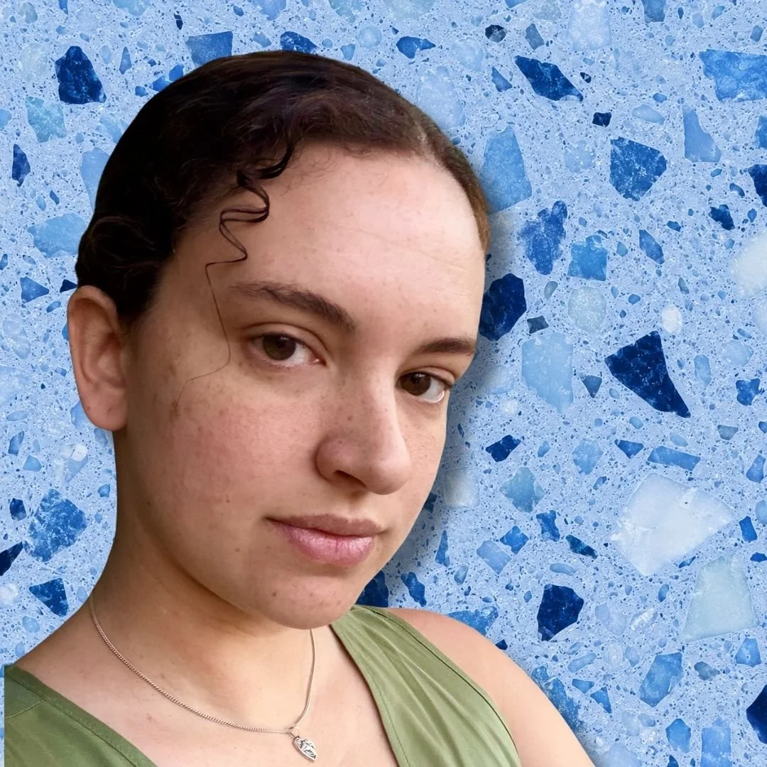 A young woman with short dark curly hair, wearing a green tank top and a silver necklace, in front of a blue terrazzo wall.