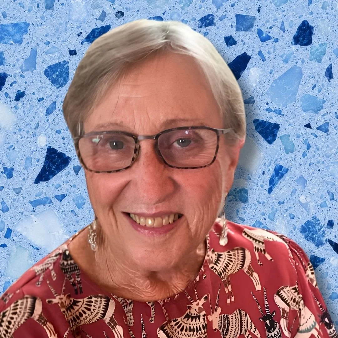 A smiling elderly woman with short gray hair and glasses, wearing a patterned blouse, in front of a blue terrazzo background.