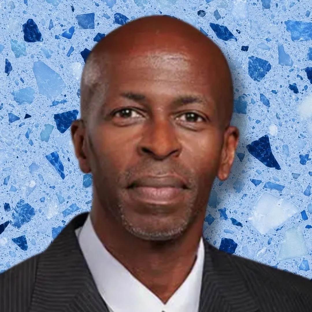 Close-up portrait of a middle-aged African American man with a bald head, wearing a black pinstripe suit and white shirt, against a blue mosaic background.