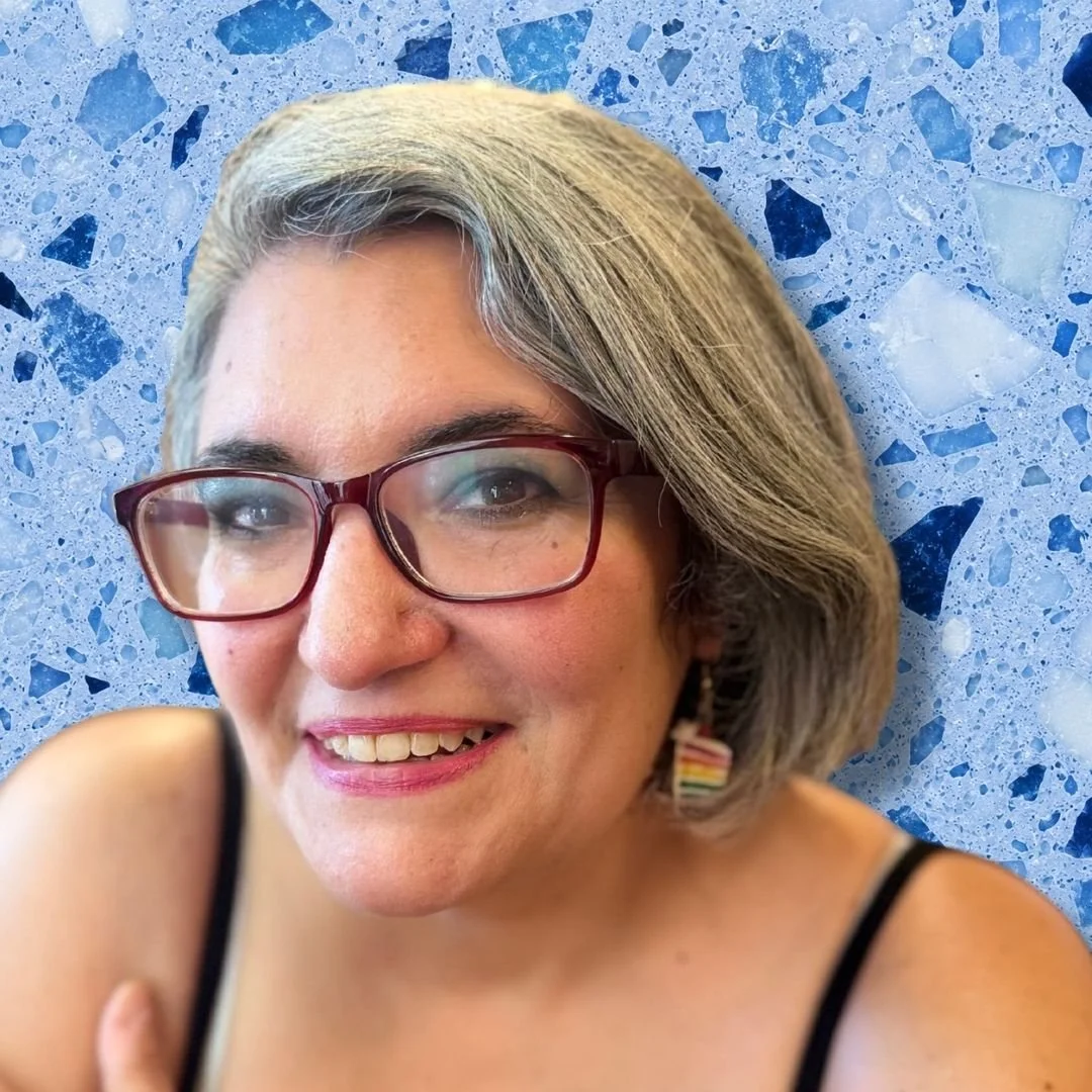 A woman with short gray hair, glasses, and earrings smiling against a blue terrazzo background.