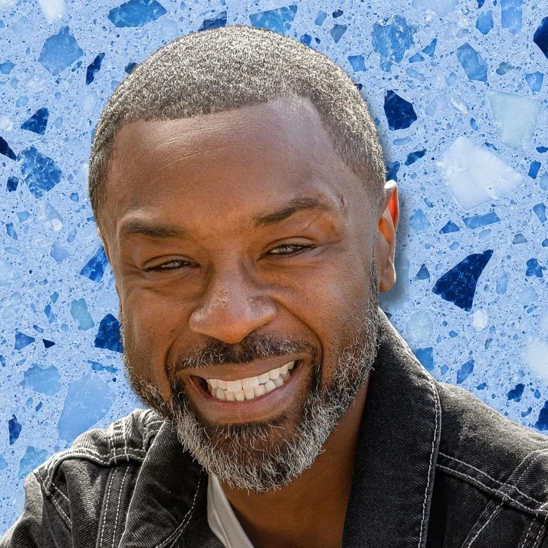 Close-up portrait of a smiling man with a gray beard and short hair, wearing a black denim jacket, with a blue terrazzo background.