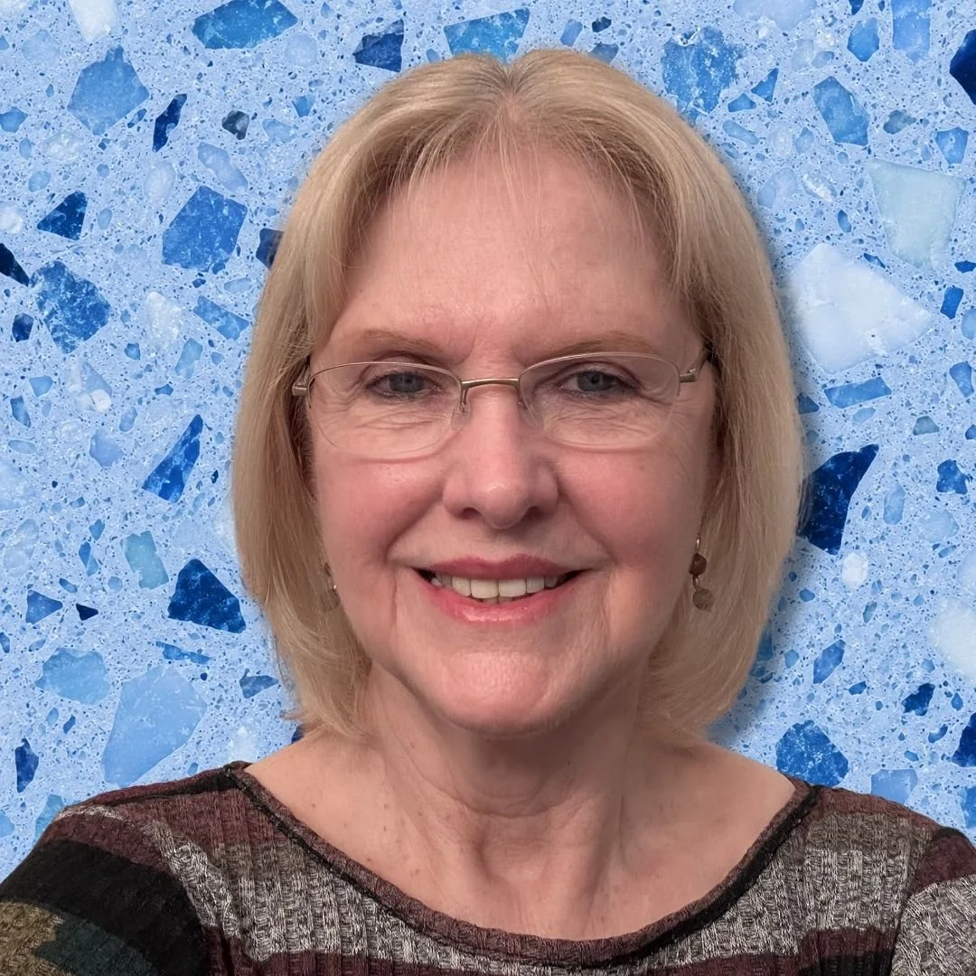 A woman with blonde hair and glasses smiling in front of a blue mosaic background.