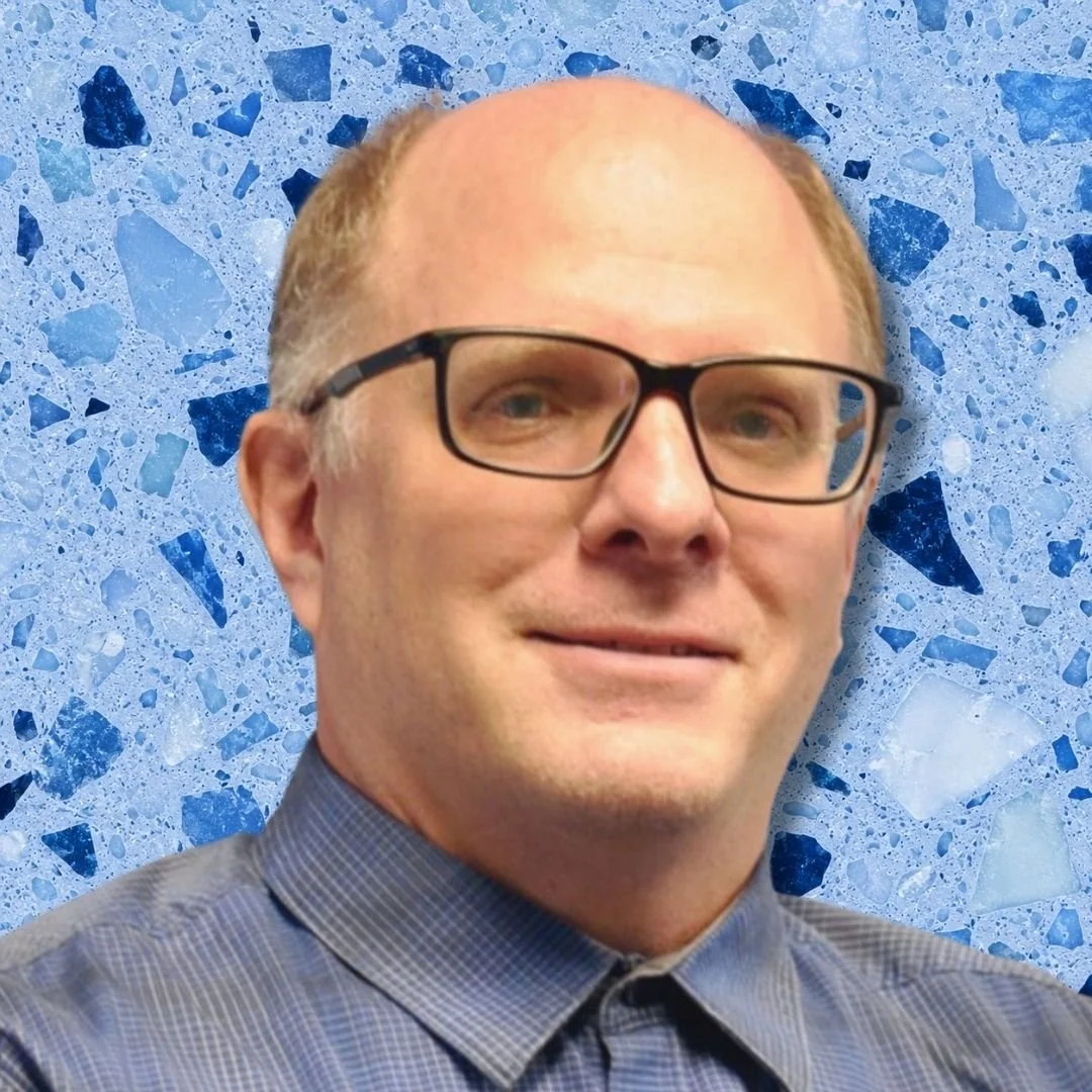 Portrait of a man with glasses and short hair, wearing a blue checkered shirt, against a blue terrazzo background.