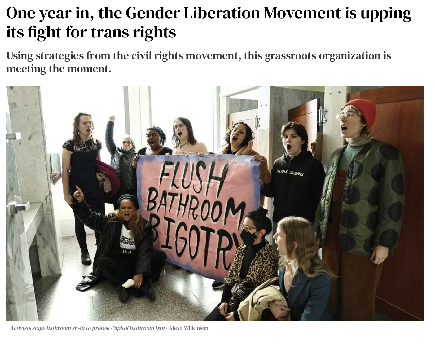 Group of activists protesting bathroom restrictions inside a building, holding a large pink sign that reads 'Flush Bathroom Bigotry', with some activists shouting and raising fists, seated and standing in front of a restroom door.