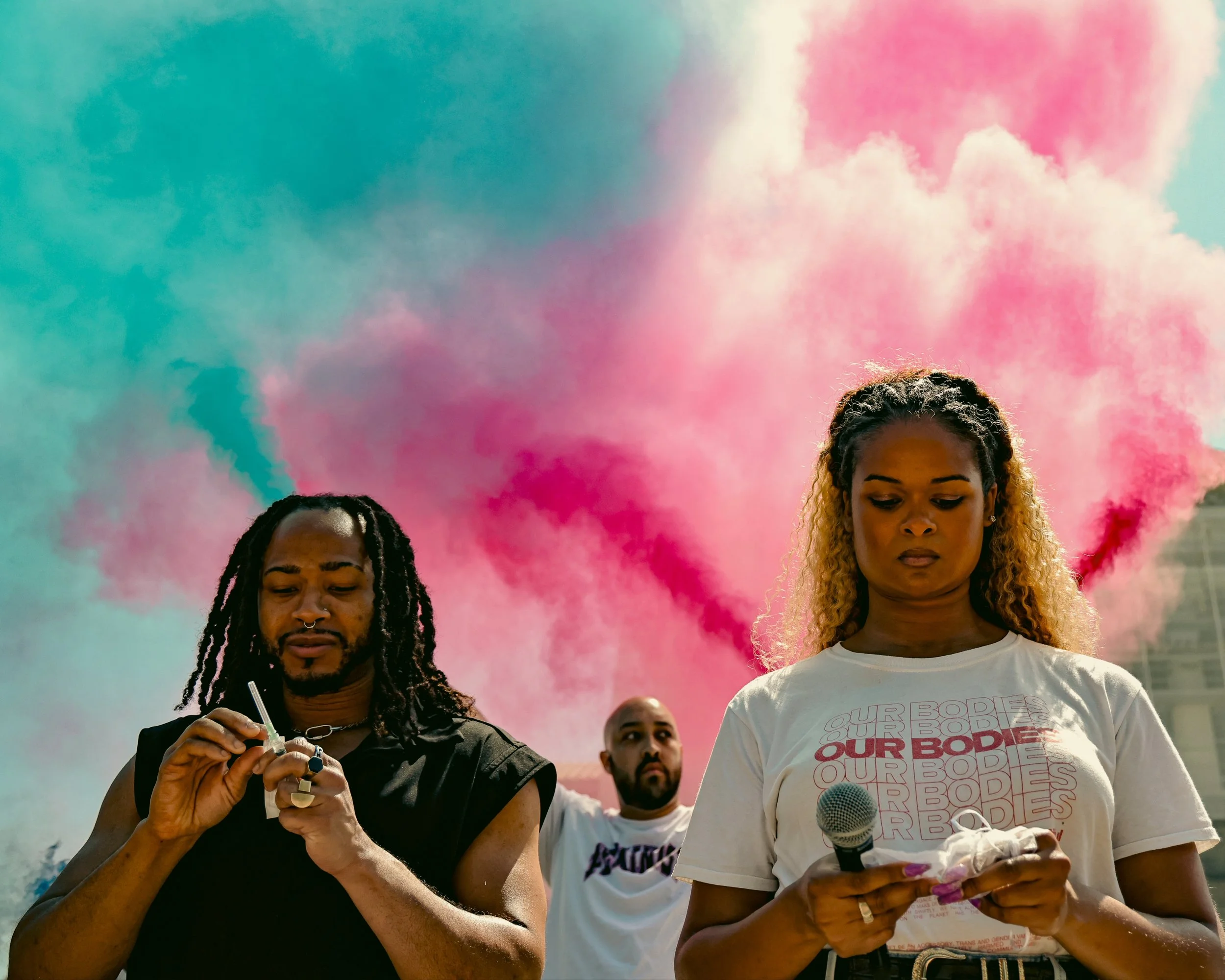 Three people standing outdoors with colorful pink and blue smoke in the background. The person on the left has dreadlocks and is holding a small object and a stick, possibly a vape or smoke device. The person in the center has a bald head and beard, with one arm raised. The person on the right has curly hair with blonde highlights, is holding a microphone and a piece of paper, and appears to be reading or preparing to speak.
