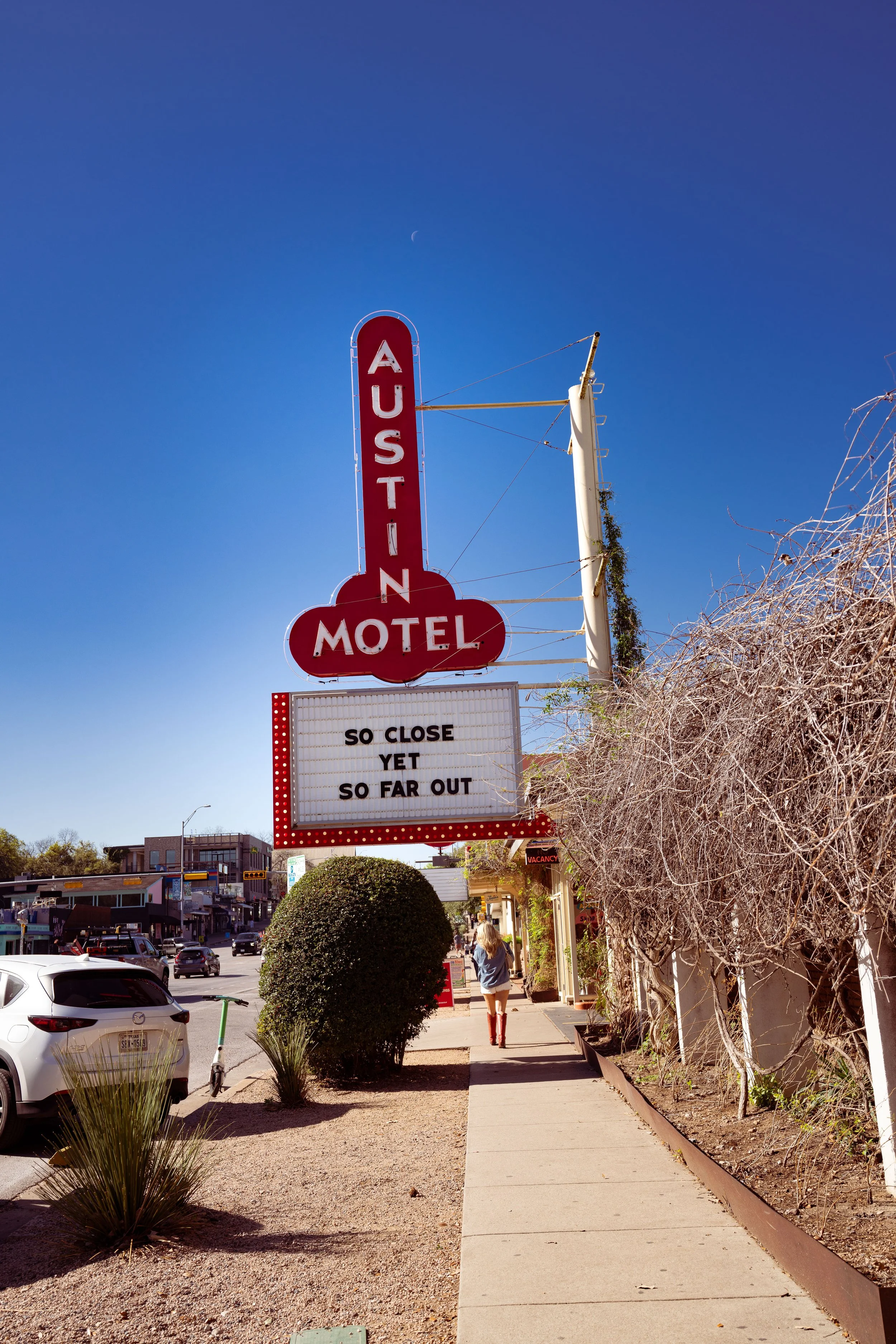 A vertical red and white sign for the Austin Motel with a humorous message on a white marquee board below, reading 'SO CLOSE YET SO FAR OUT'. The scene is a sunny street with a woman walking, cars parked along the sidewalk, and desert plants and bushes.
