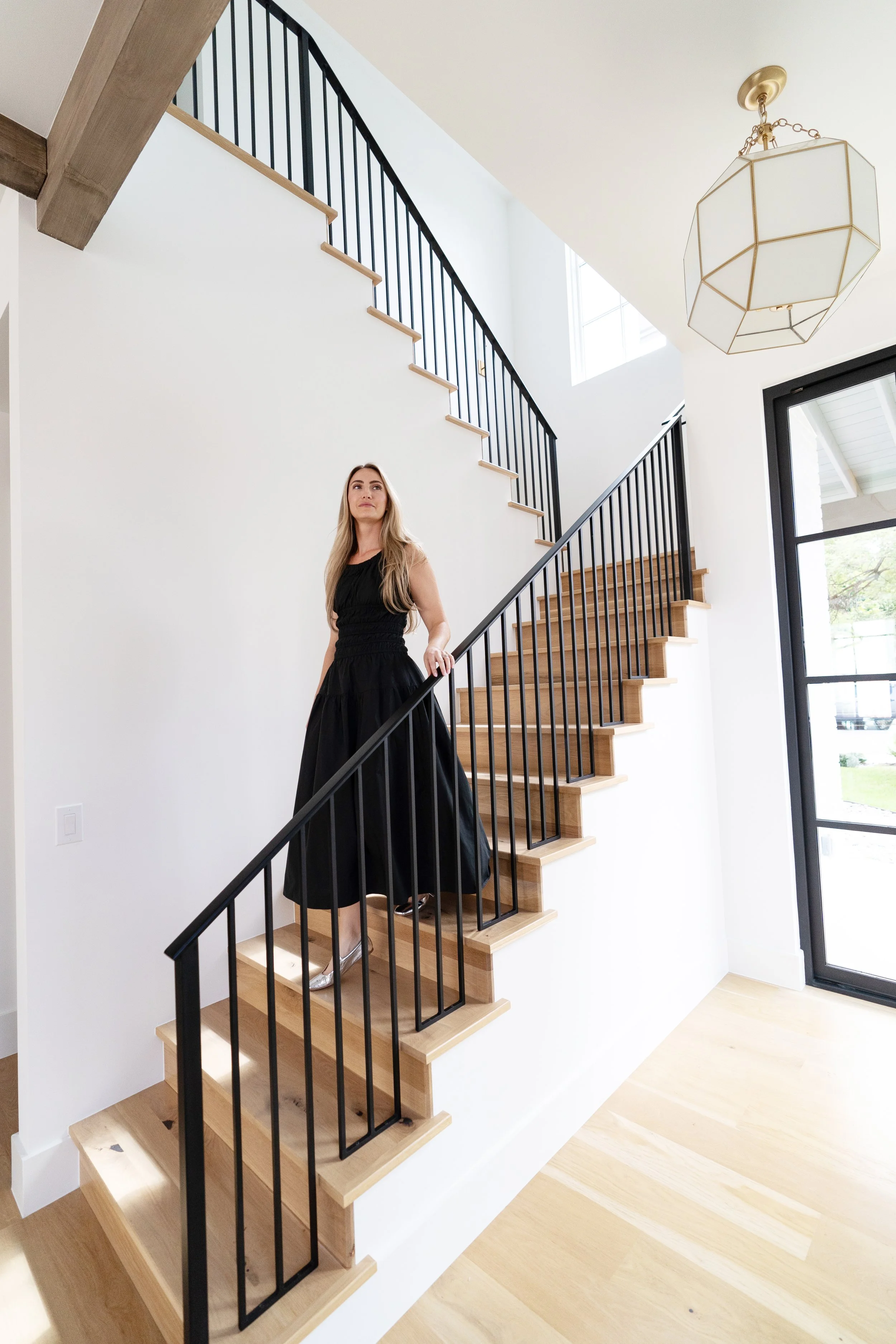 Woman in black dress standing on wooden staircase with black railings inside a modern home with large windows and a geometric light fixture.