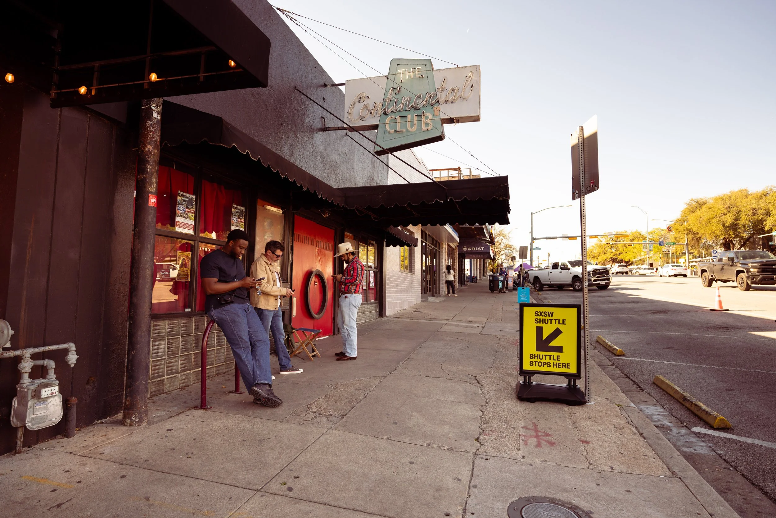 People waiting at the sidewalk outside a bar called The Continental Club, with a yellow sign indicating a shuttle stop for SXSW.