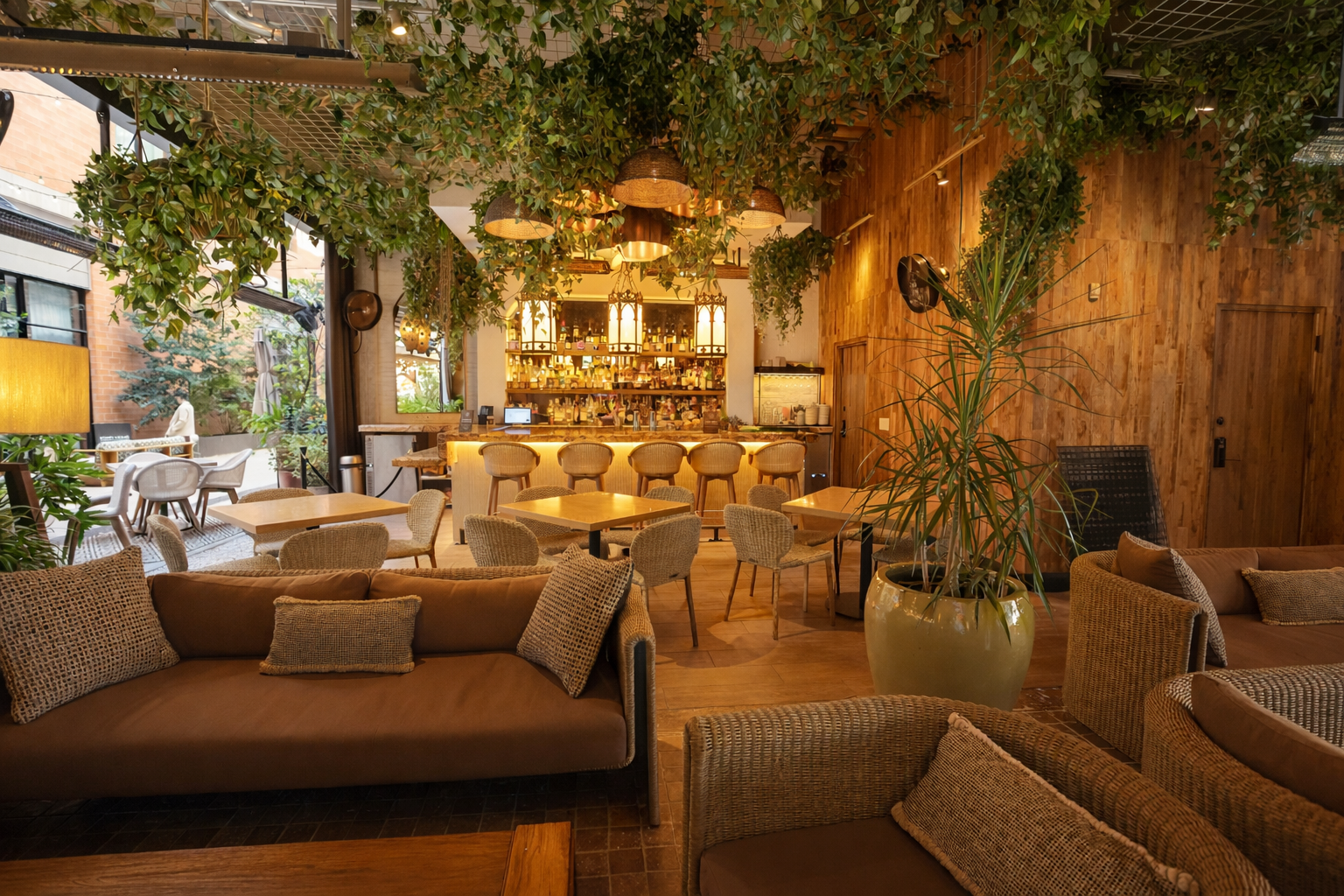 A cozy restaurant interior featuring wooden walls, seating with cushions, a large potted plant, and a bar area with hanging lamps and greenery overhead.