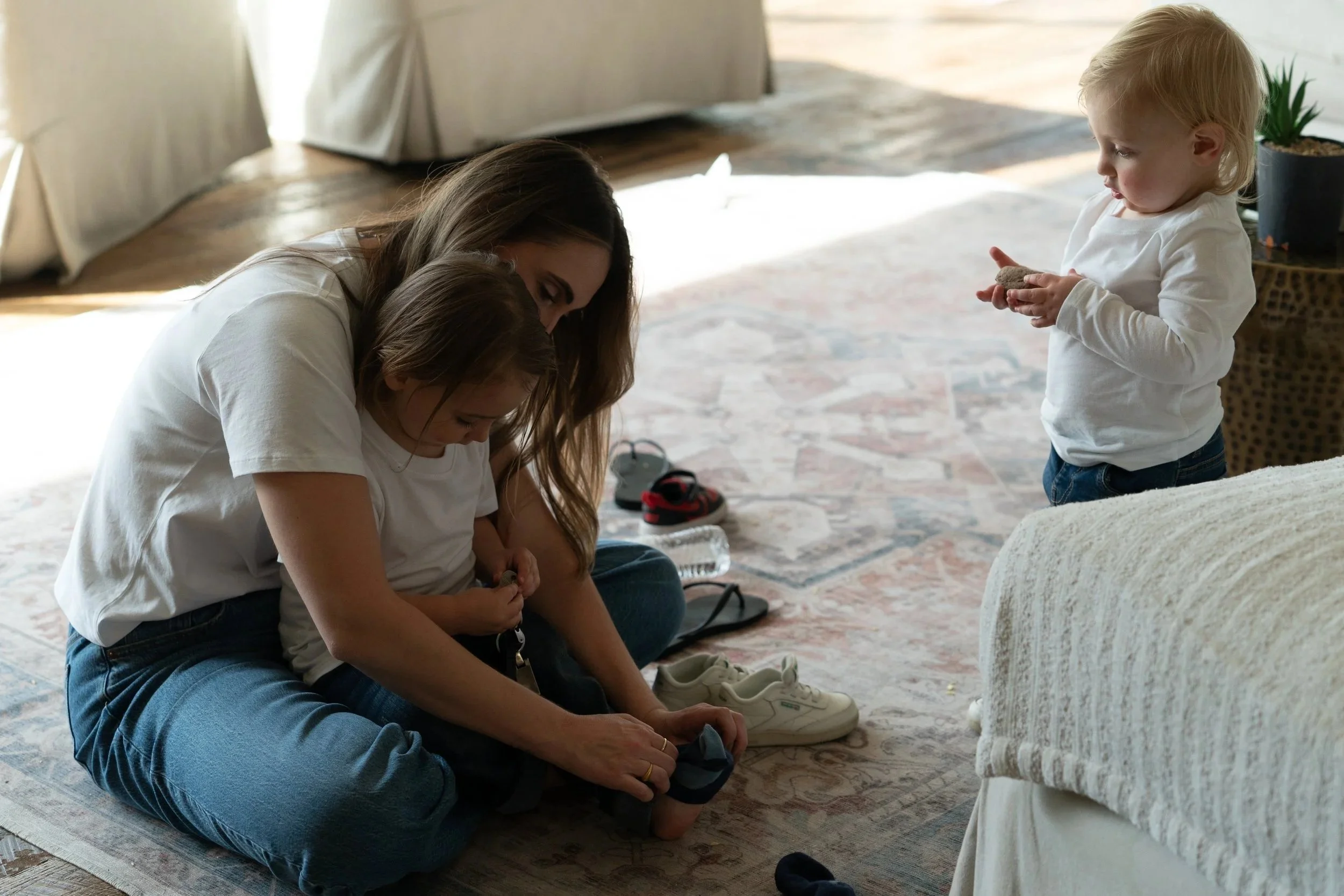 A woman and a child sitting on the floor, with the woman helping the child put on shoes. A young girl stands nearby, holding a small object, and there are shoes and a water bottle on the floor.