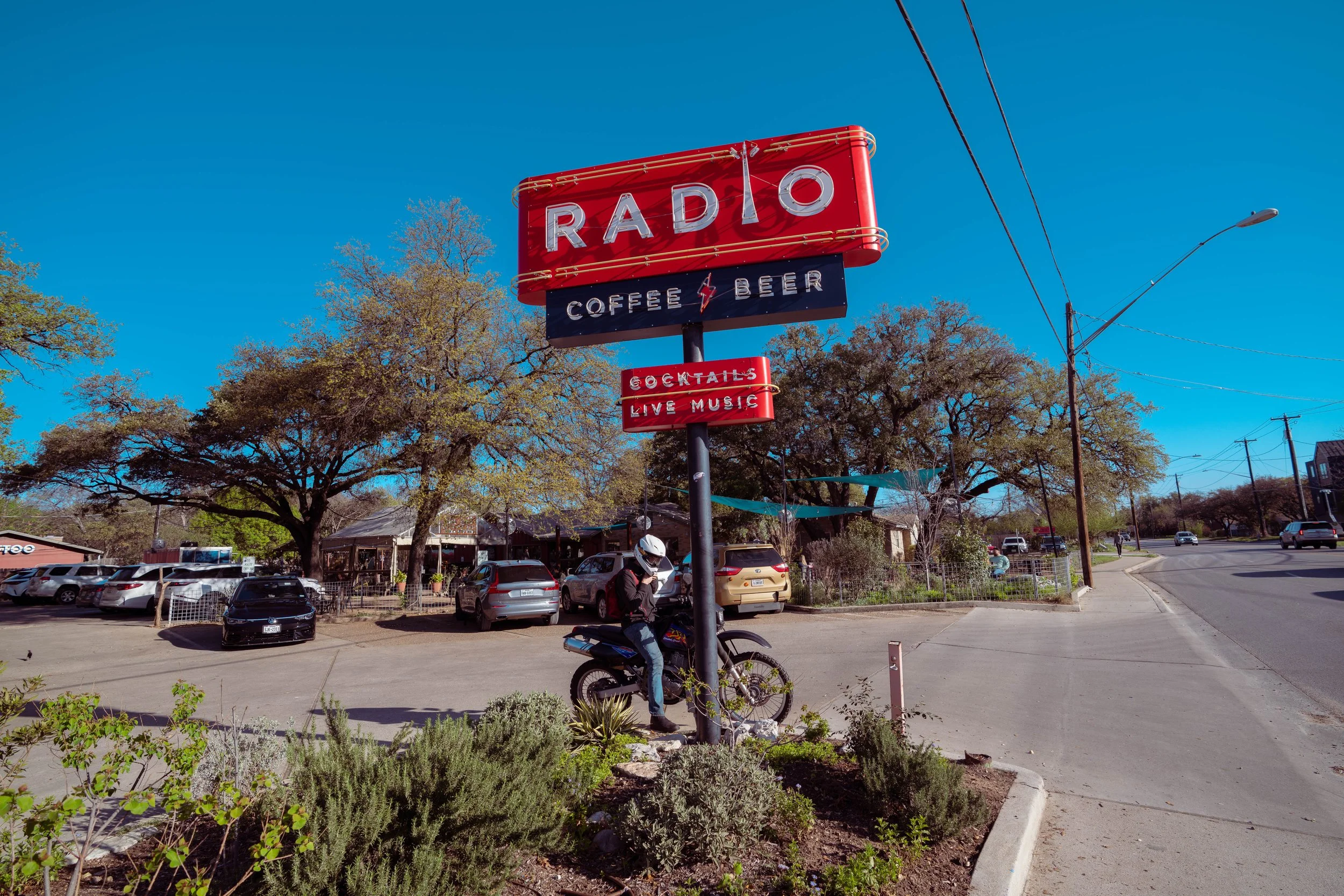 A street view with a large red and white Radio signboard advertising coffee, beer, cocktails, and live music. A person wearing a helmet is sitting on a motorcycle next to the sign. The background features trees, parked cars, and a clear blue sky.