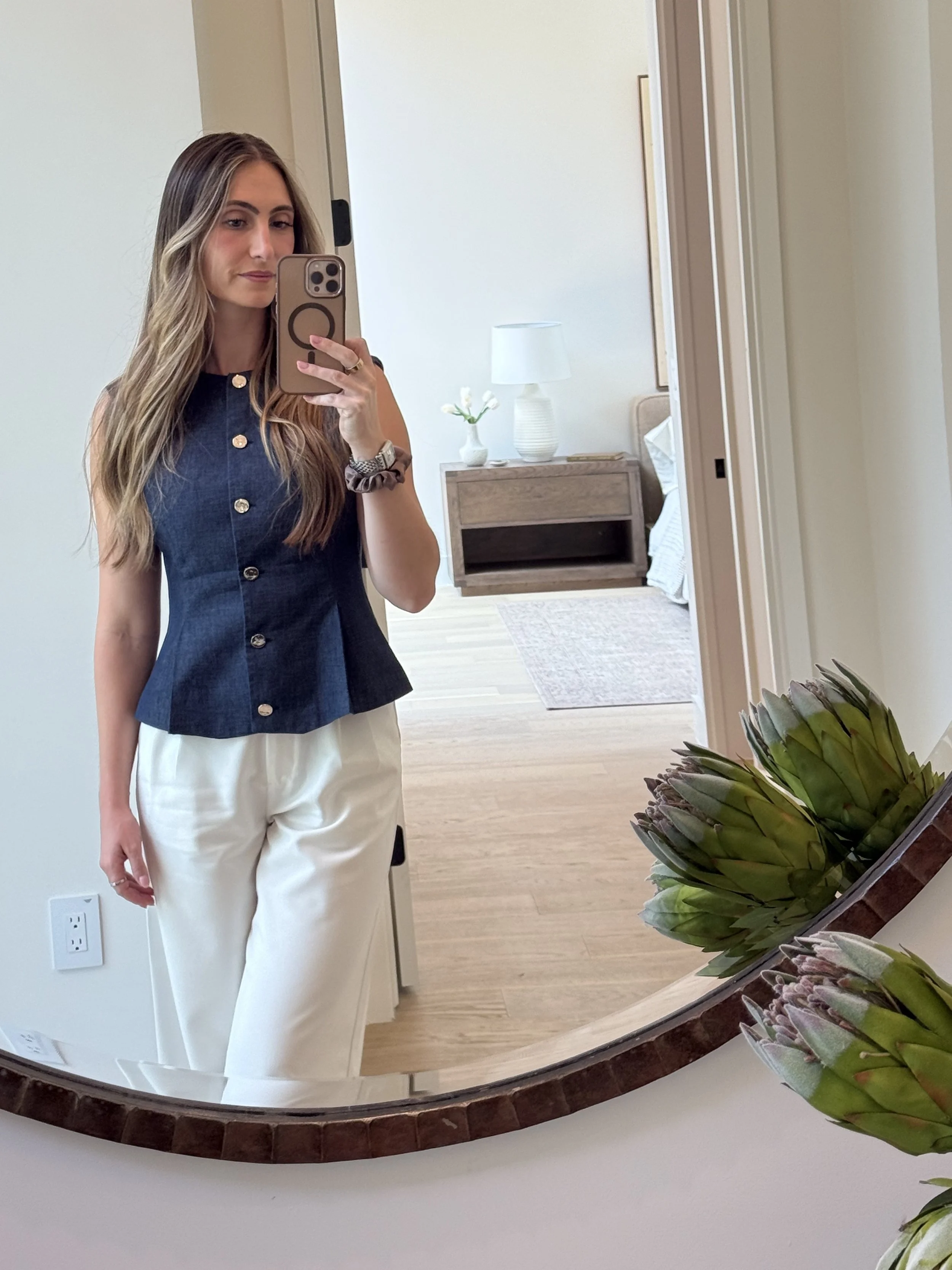 Woman taking a mirror selfie in a bedroom, wearing a sleeveless denim top and white pants, with a wooden nightstand, lamp, and flowers in the background.