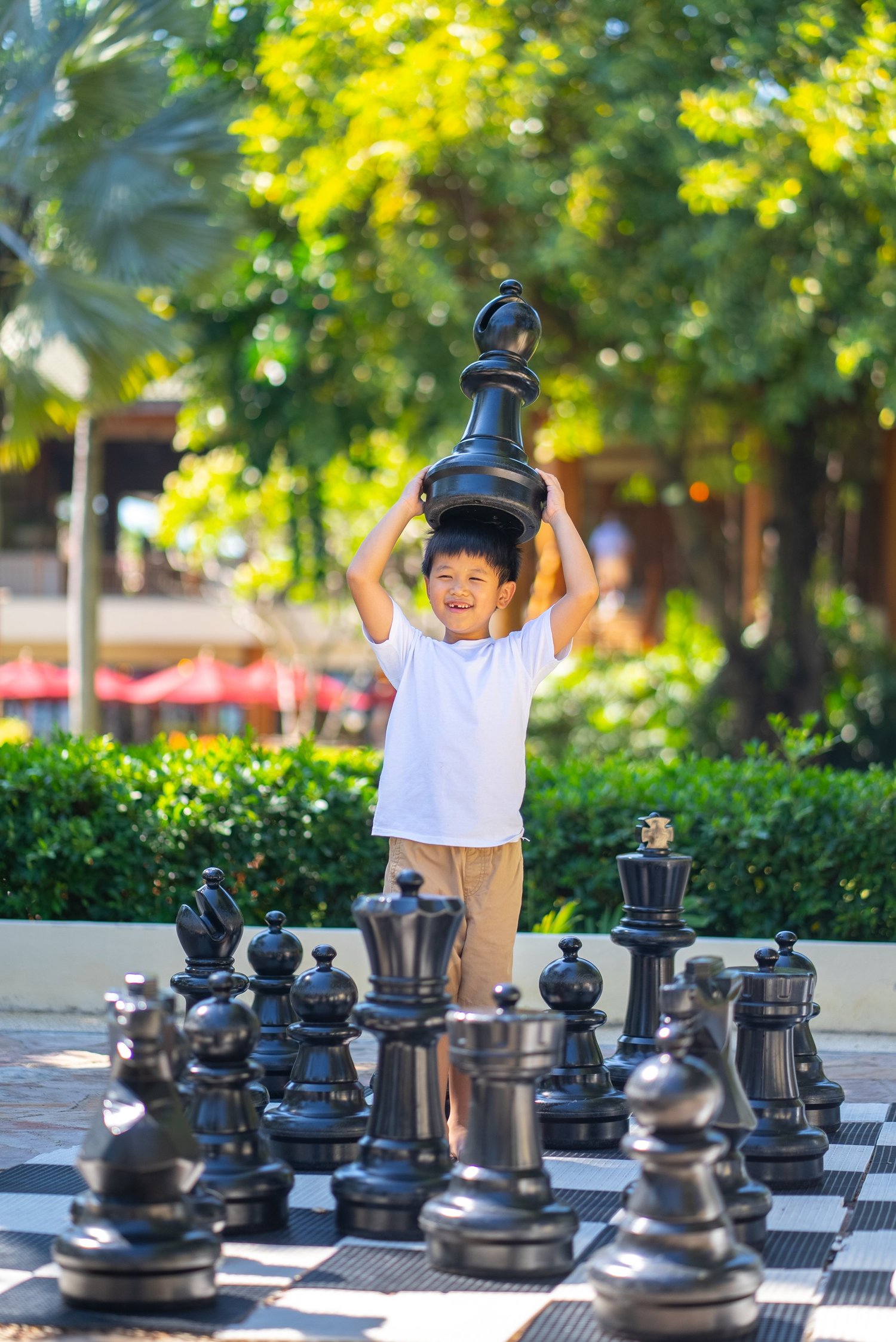 A young boy grinning as he lifts a giant chess piece above his head on an outdoor chessboard