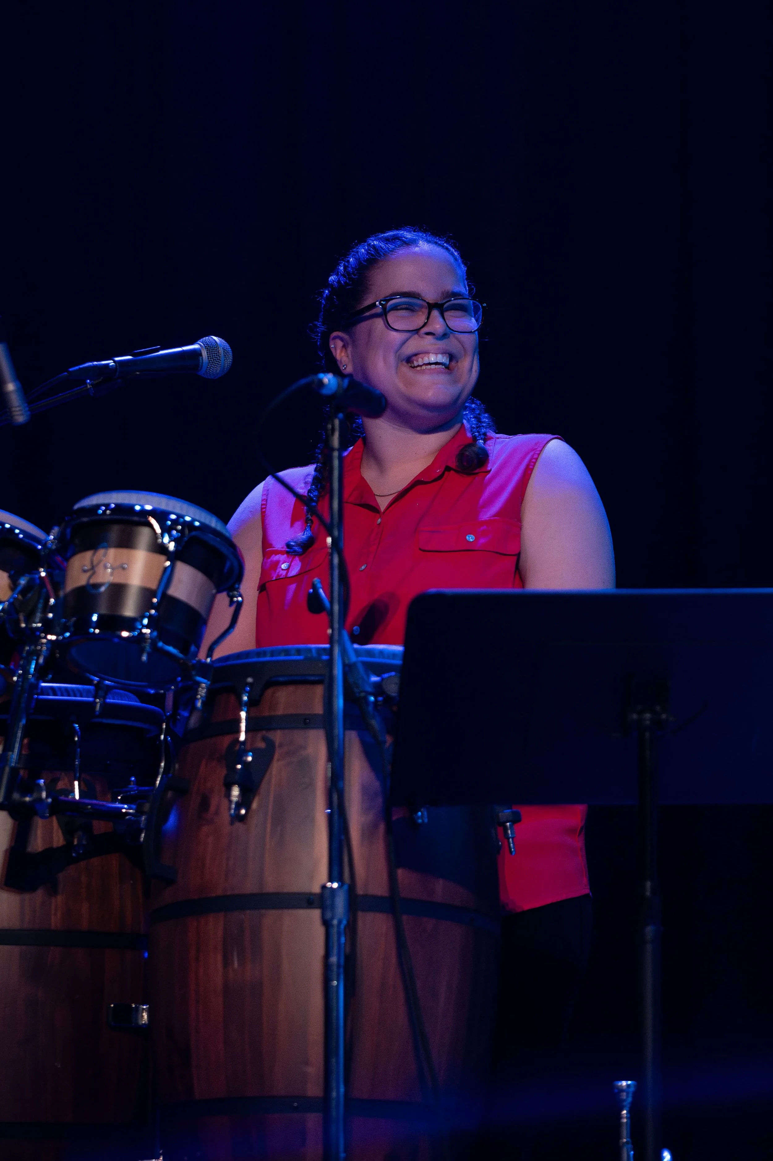 A woman with glasses, wearing a red sleeveless shirt, smiling while standing behind percussion instruments on stage. Corporate party entertainment in Lancaster PA with Seraphina Soul live band.