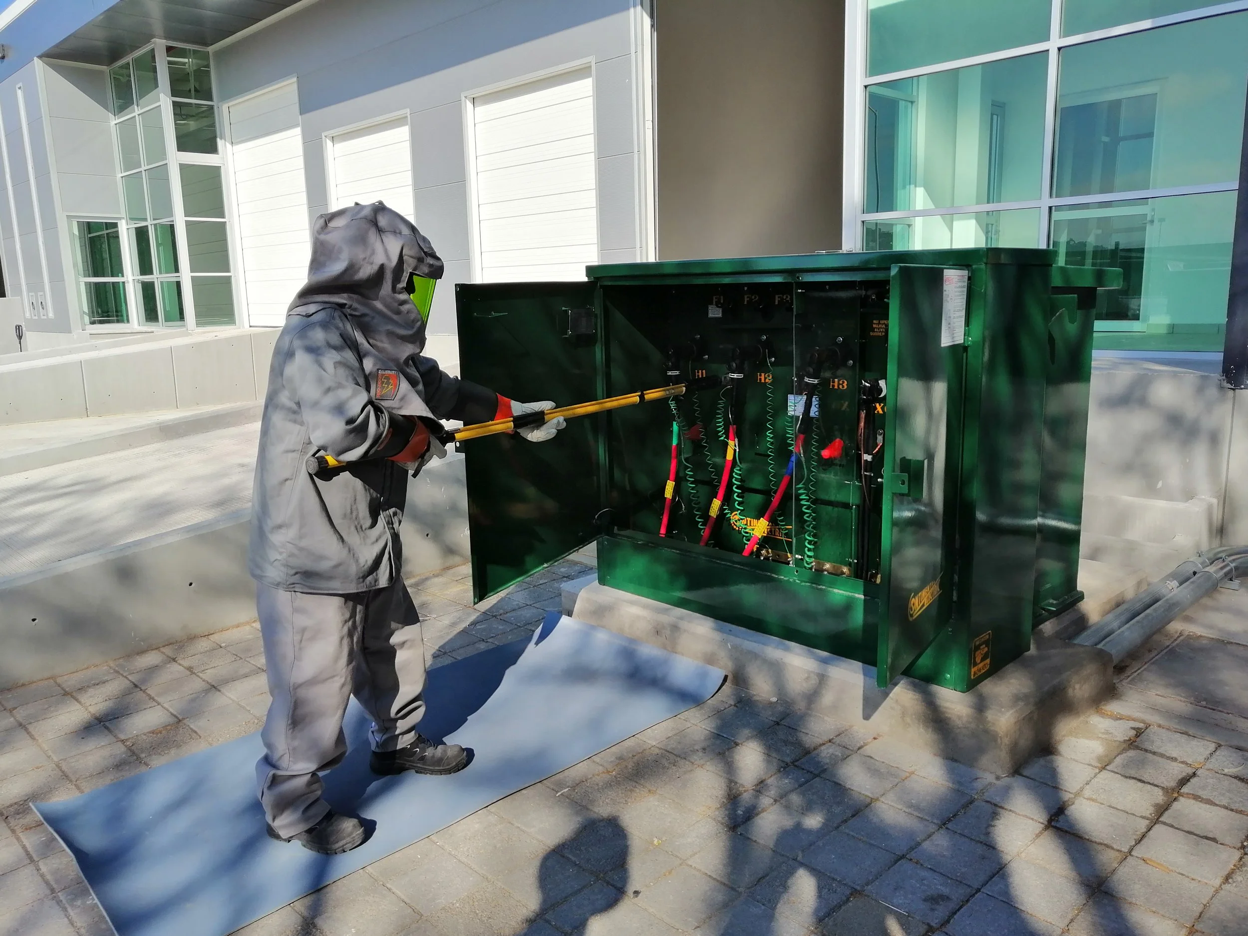 Técnico trabajando en una caja de componentes eléctricos o de energía con equipo de protección y herramienta de equipo, en un entorno exterior con edificio de fondo.