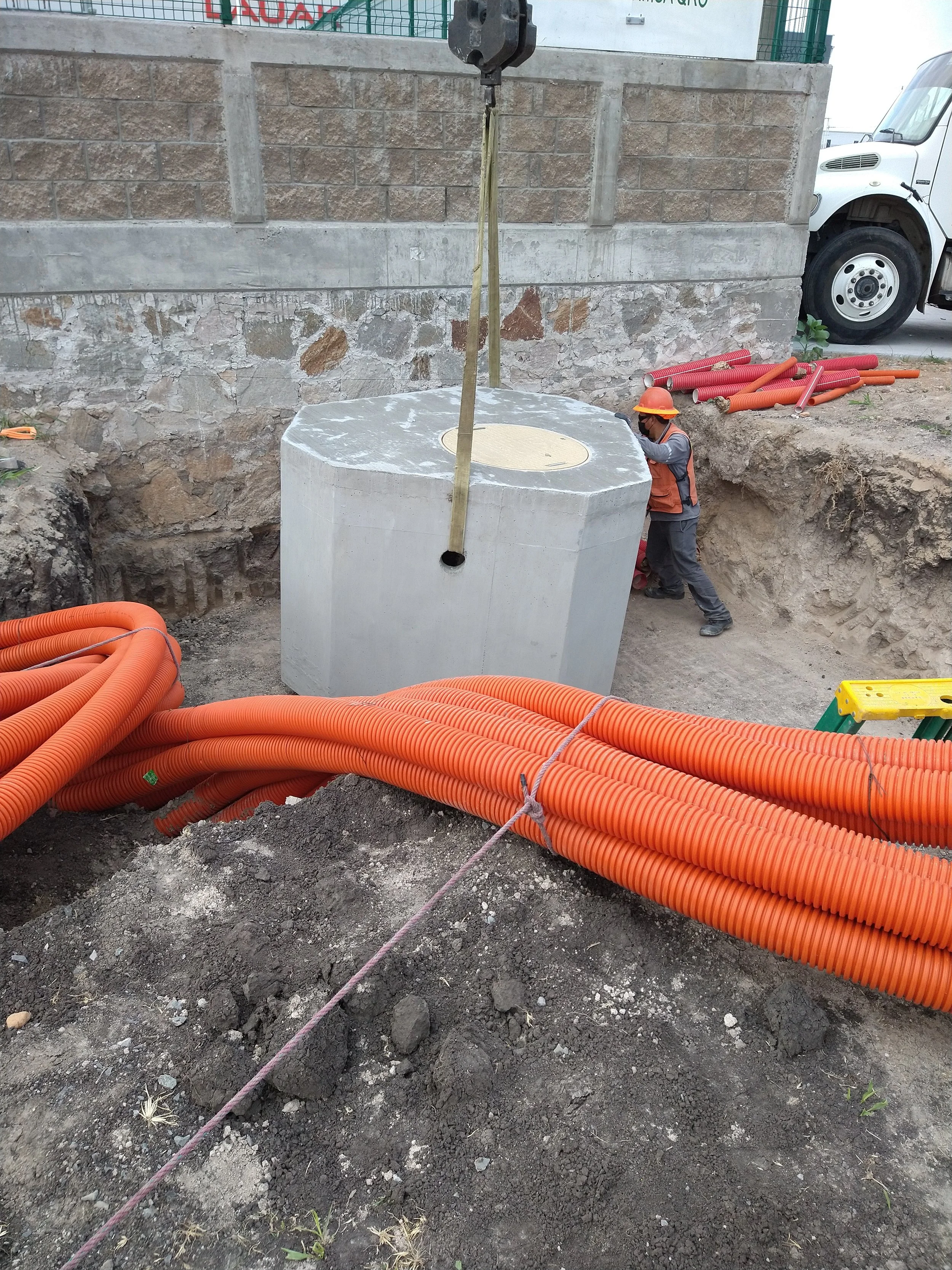 Trabajador de construcción usando casco y cinturón de seguridad trabajando con una estructura de concreto y múltiples mangueras de plástico naranja en un sitio de excavación.