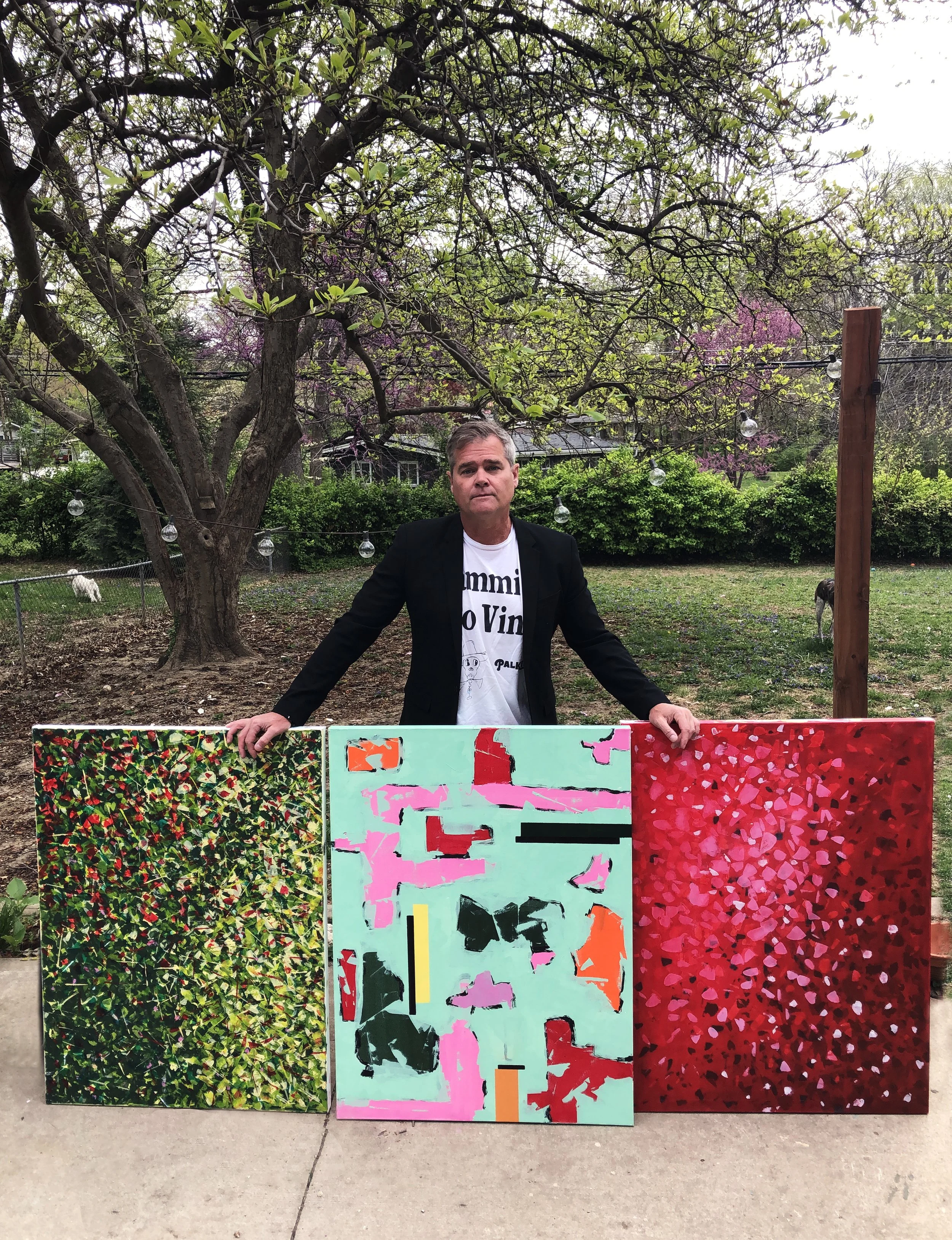 A man standing behind three colorful abstract paintings outdoors in a garden, with trees and blooming flowers in the background.