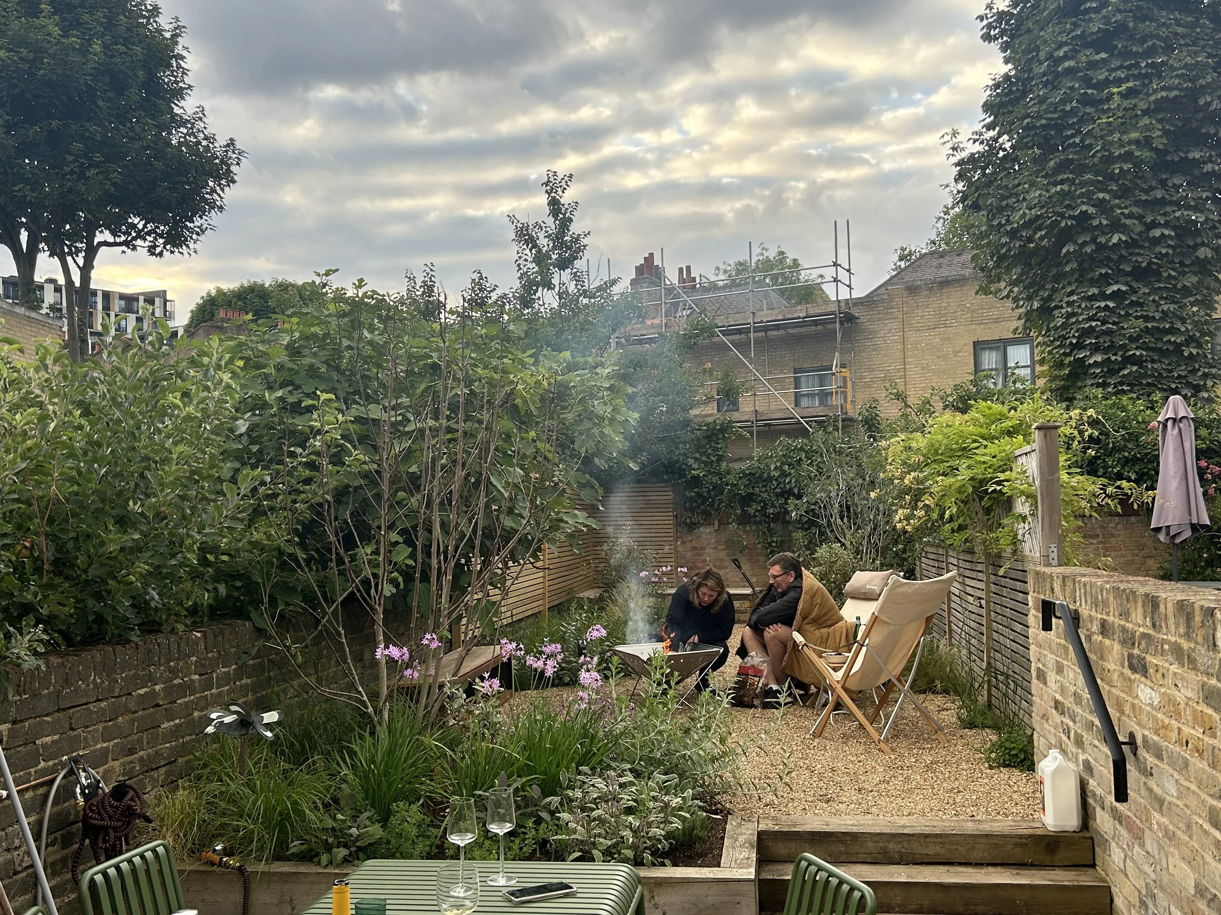 A backyard with lush green plants, trees, and pink flowers. Two people are sitting and chatting by a small fire. There are chairs, glasses on the table, and a garden fence with a brick wall.