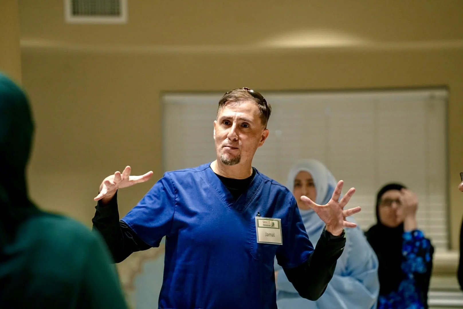 A man in blue scrubs with a name tag that says 'Jamal' is speaking to a group of people, some of whom are wearing headscarves, in a room with beige walls and closed blinds.