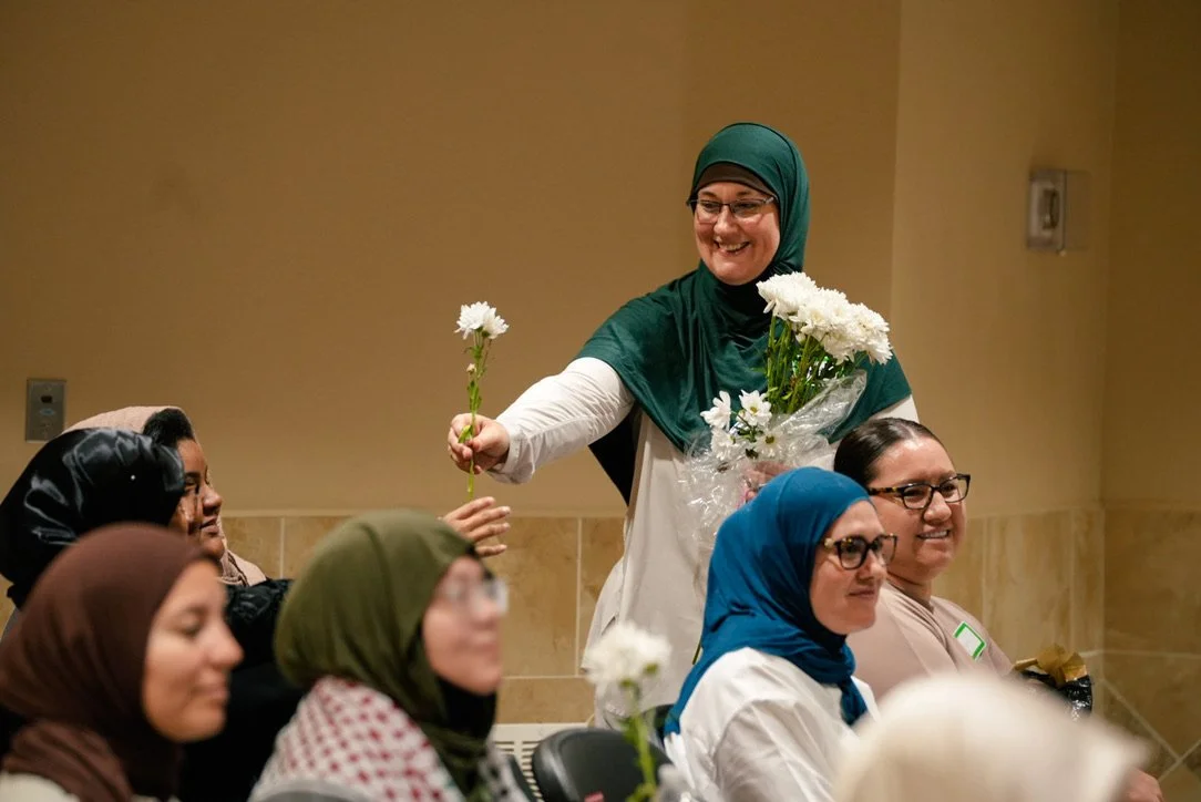 Women, some wearing headscarves, sitting in a room while one woman in a green headscarf and glasses offers a white flower to another woman.
