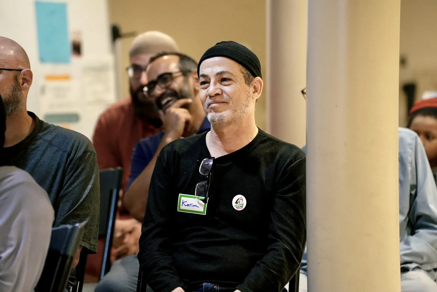 A group of people sitting, with one person in the center wearing a black beanie, black shirt, and glasses hanging from the shirt collar, smiling and looking to the side.