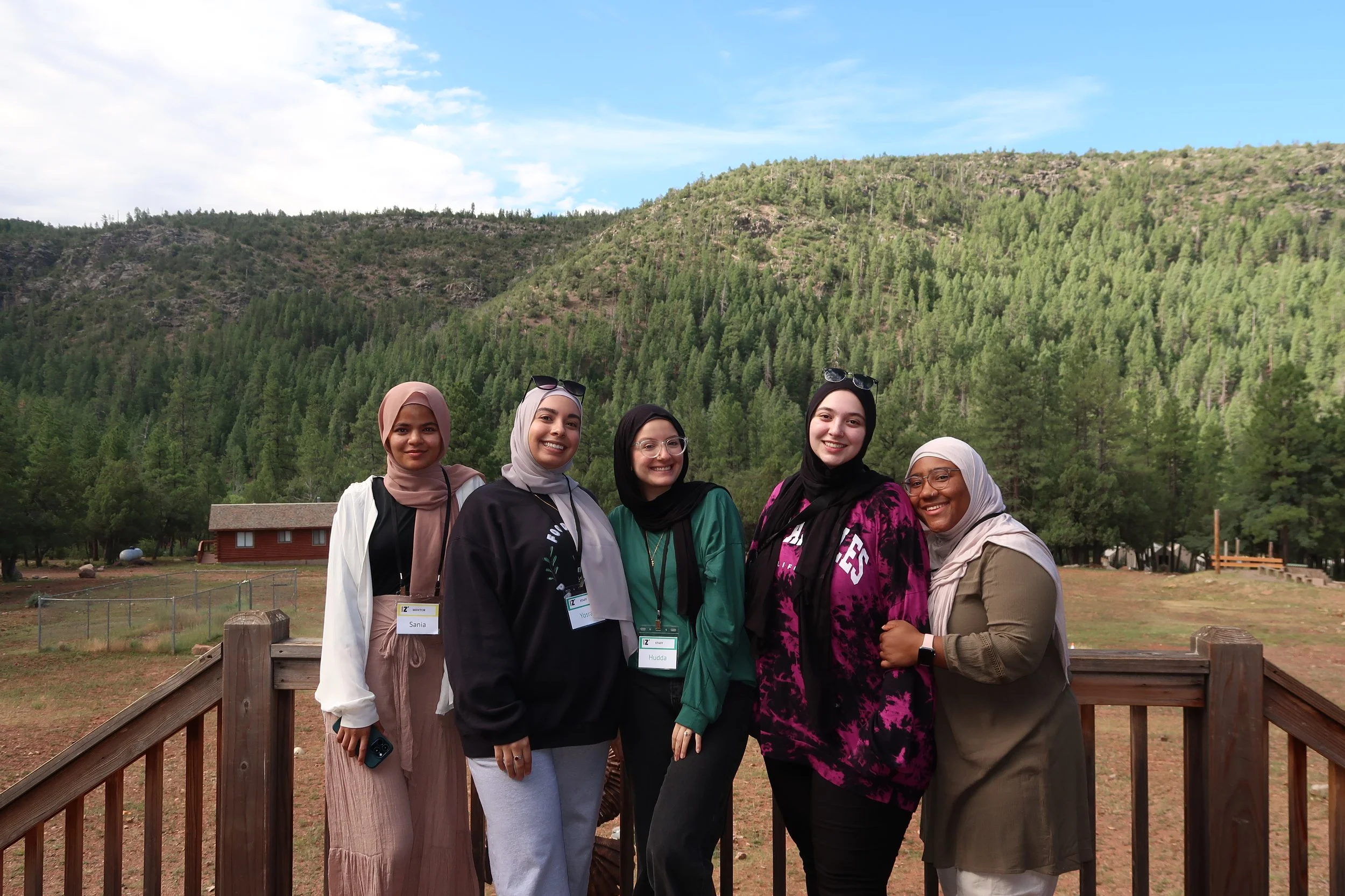 Five women wearing hijabs and casual clothing pose together outdoors in front of a forested mountain landscape, smiling at the camera.