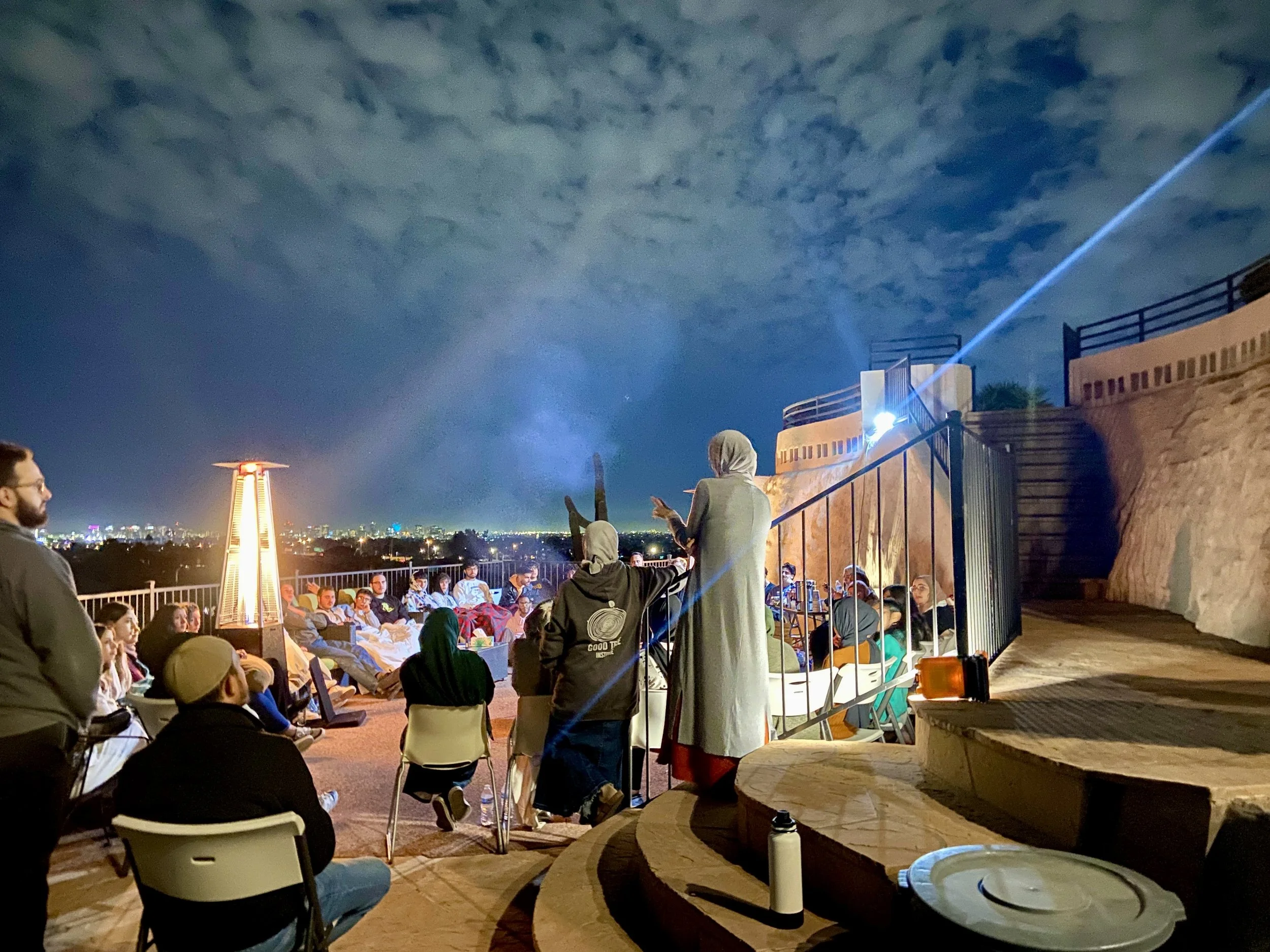 People sitting outdoors at night, gathered for a presentation or event, with a person standing and speaking in front of the group. The sky is cloudy and illuminated by city lights in the distance, with a bright spotlight and an outdoor heater visible