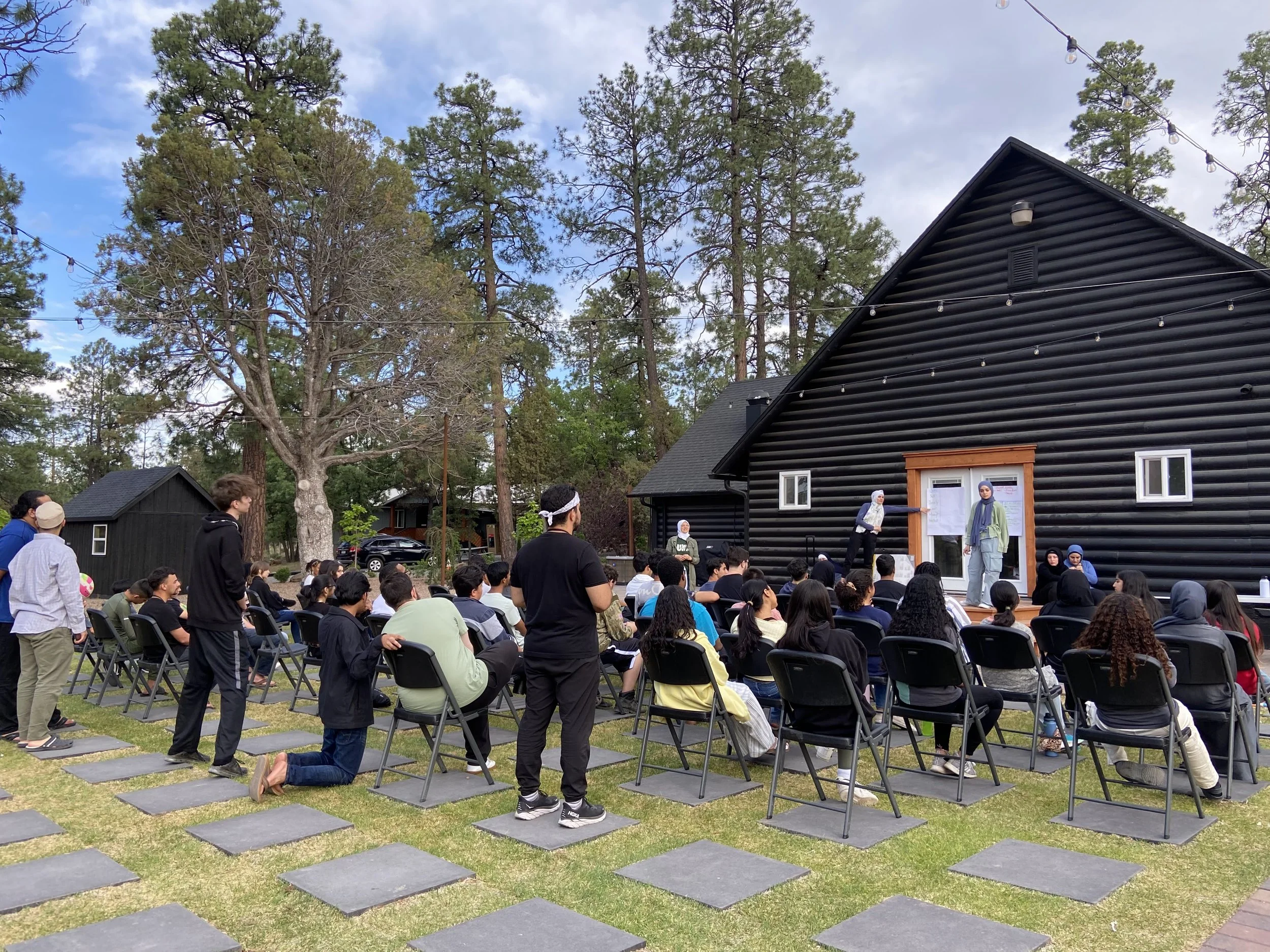 Outdoor gathering with people seated in chairs facing a small stage in front of a black wood-paneled building, with a large tree and tall pine trees in the background.
