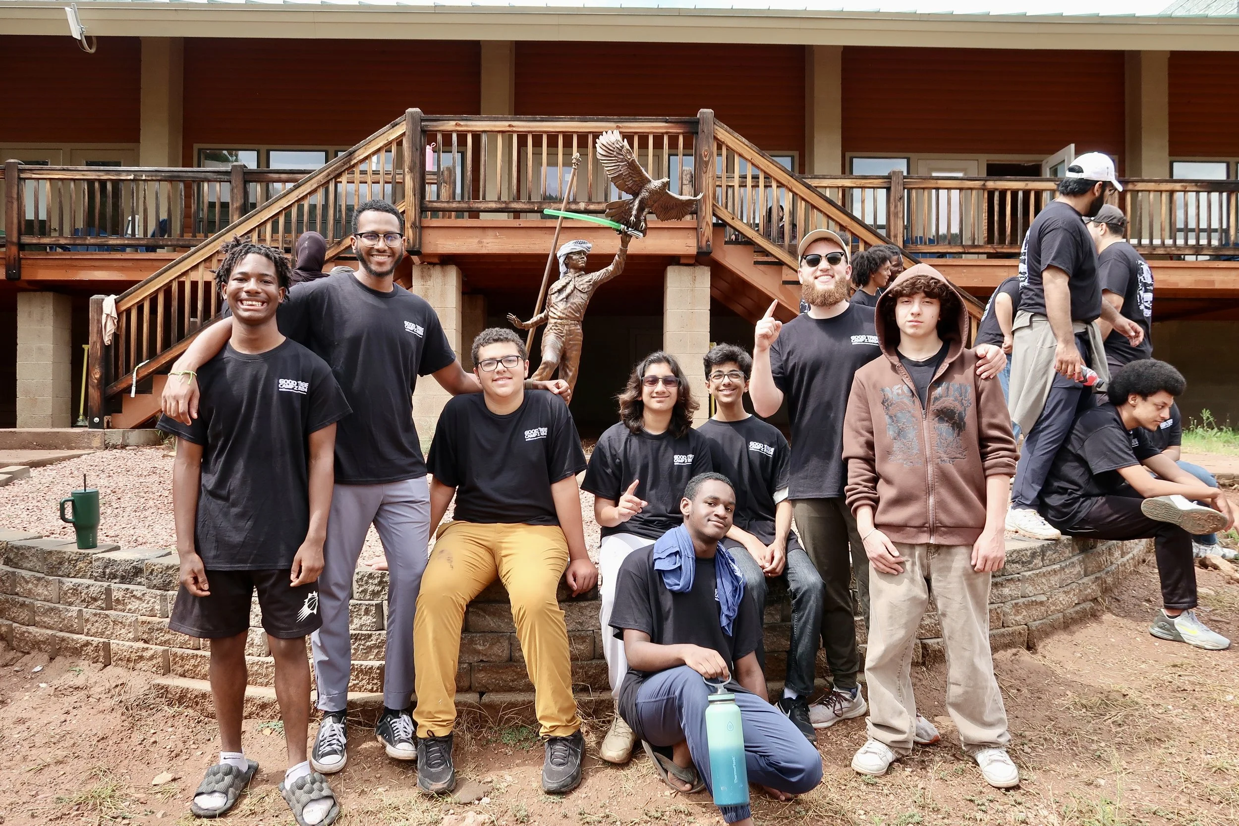 Group of young people posing outdoors in front of a wooden staircase and statue of an eagle, some wearing black t-shirts, in a casual setting.