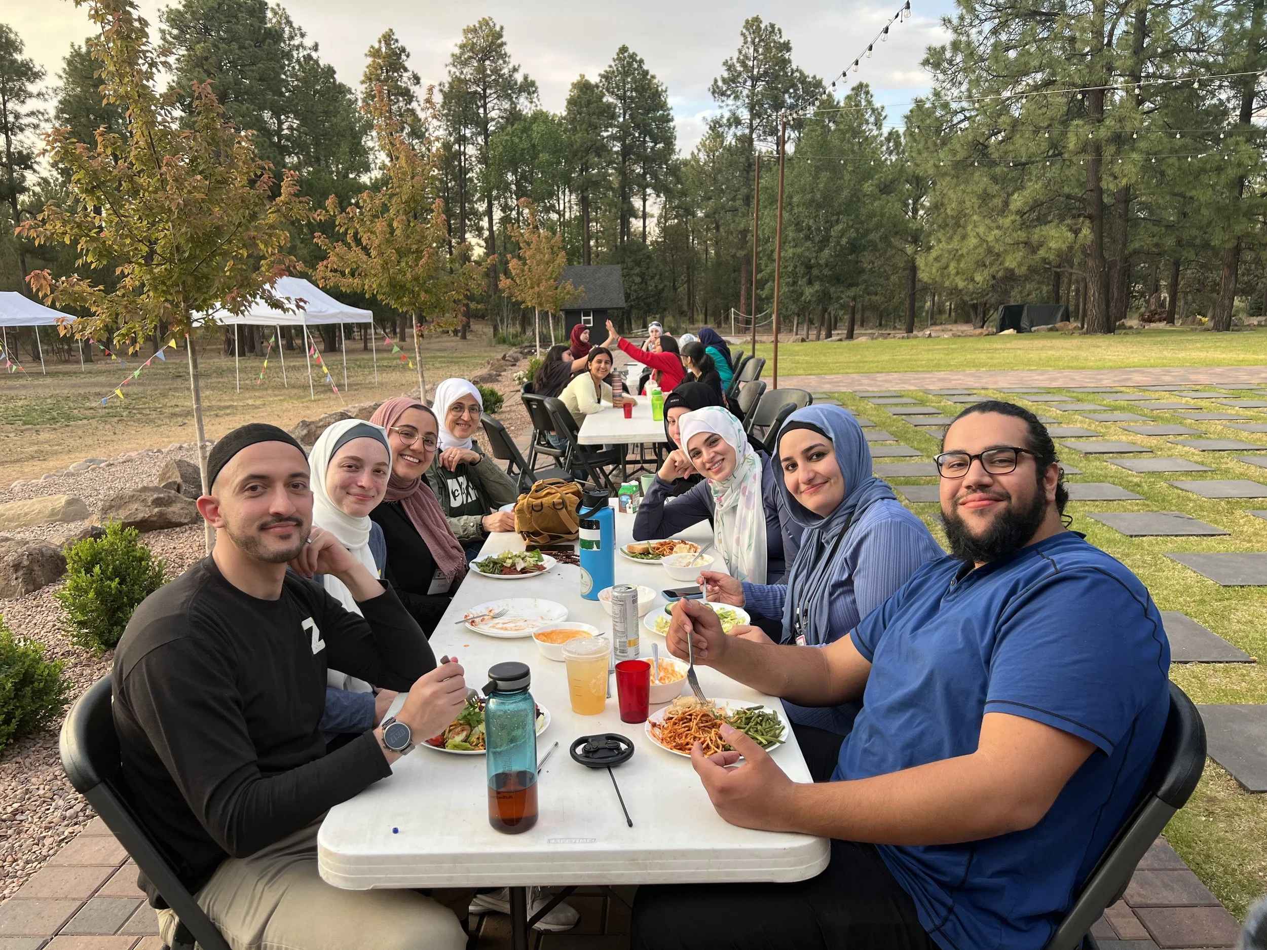 A group of people sitting at a long outdoor table enjoying a meal at a gathering in a park with trees and tents in the background.