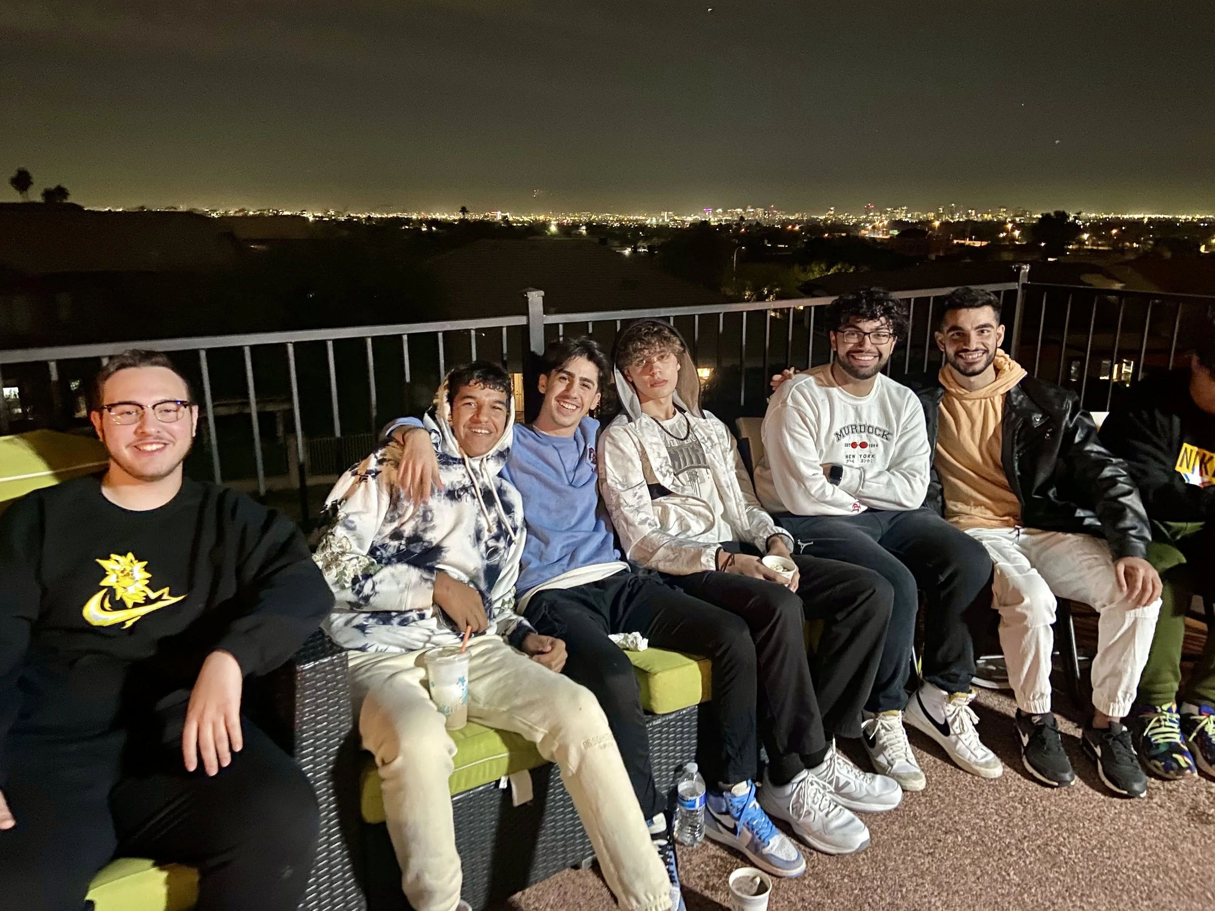 Group of seven young men sitting on a patio at night with a city skyline in the background. They are smiling and dressed casually.