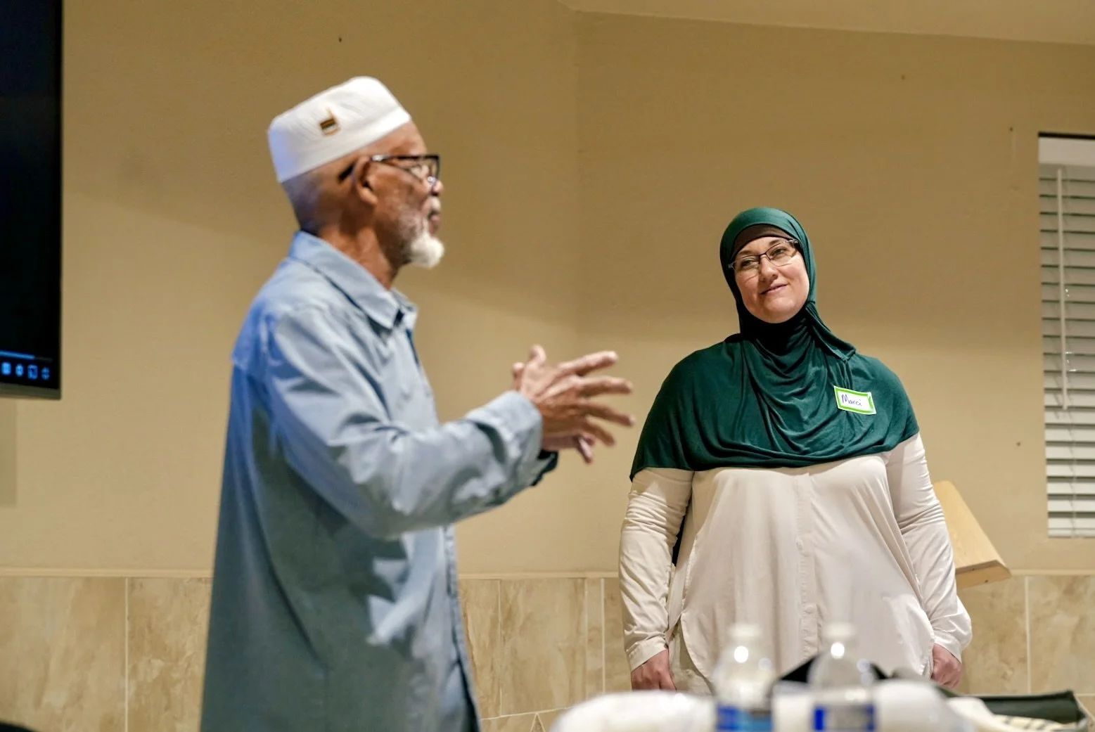 An older man with glasses, a white beard, and wearing a white cap and light blue shirt is speaking to a woman wearing a green hijab and beige top. They are indoors, possibly at a community or religious gathering.